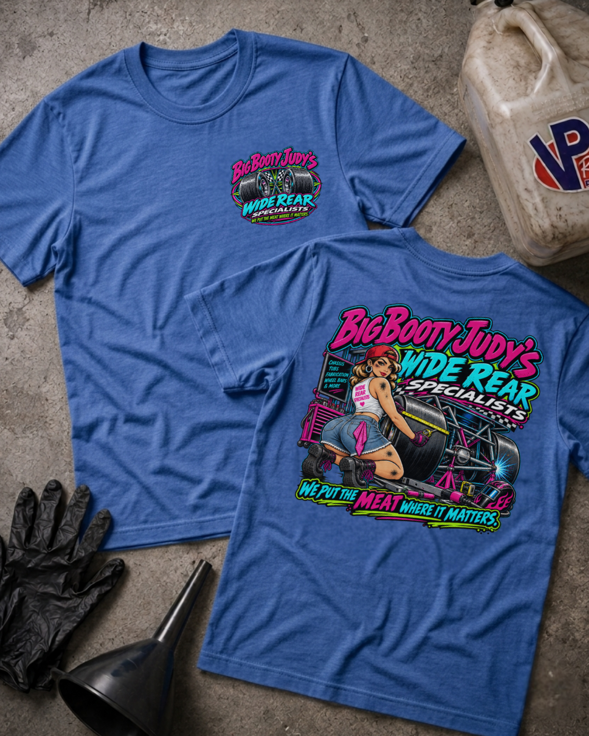 Two heather blue graphic T-shirts laid flat on a concrete surface, showing the front and back designs for 'Big Booty Judy's Wide Rear Specialists'. The front features a small pink and green logo on the left chest, while the back displays a large, colorful pin-up style graphic with a woman and the brand name.