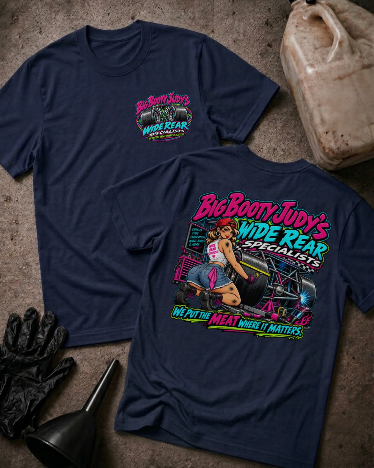 Two navy blue graphic T-shirts laid flat on a textured concrete-like surface, with a black glove, funnel, and plastic jug as props. The front shirt shows a small pink and teal chest graphic reading 'Big Boott Judy's Wide Rear Specialists,' and the back shirt shows a large colorful retro-style graphic with the same brand text and additional wording.