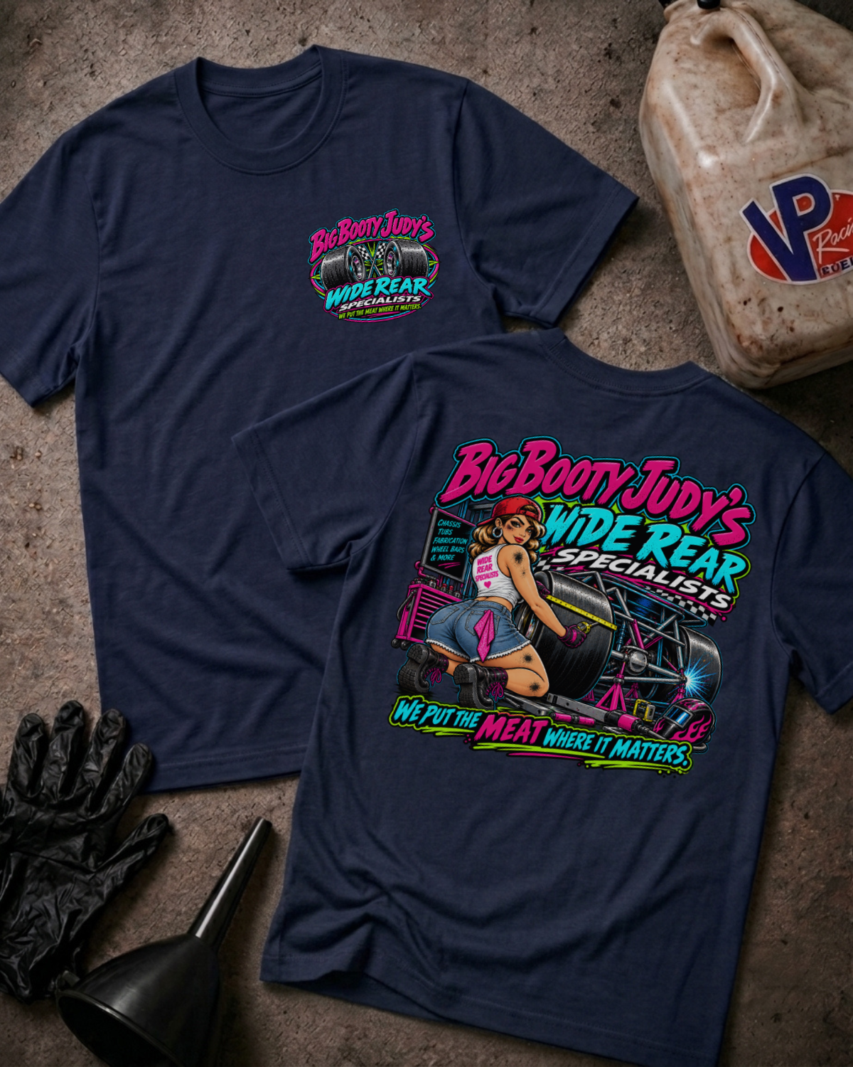 Two navy graphic T-shirts laid flat on a concrete surface, showing the front and back designs for 'Big Booty Judy's Wide Rear Specialists'. The front features a small pink and green logo on the left chest, while the back displays a large, colorful pin-up style graphic with a woman and the brand name.