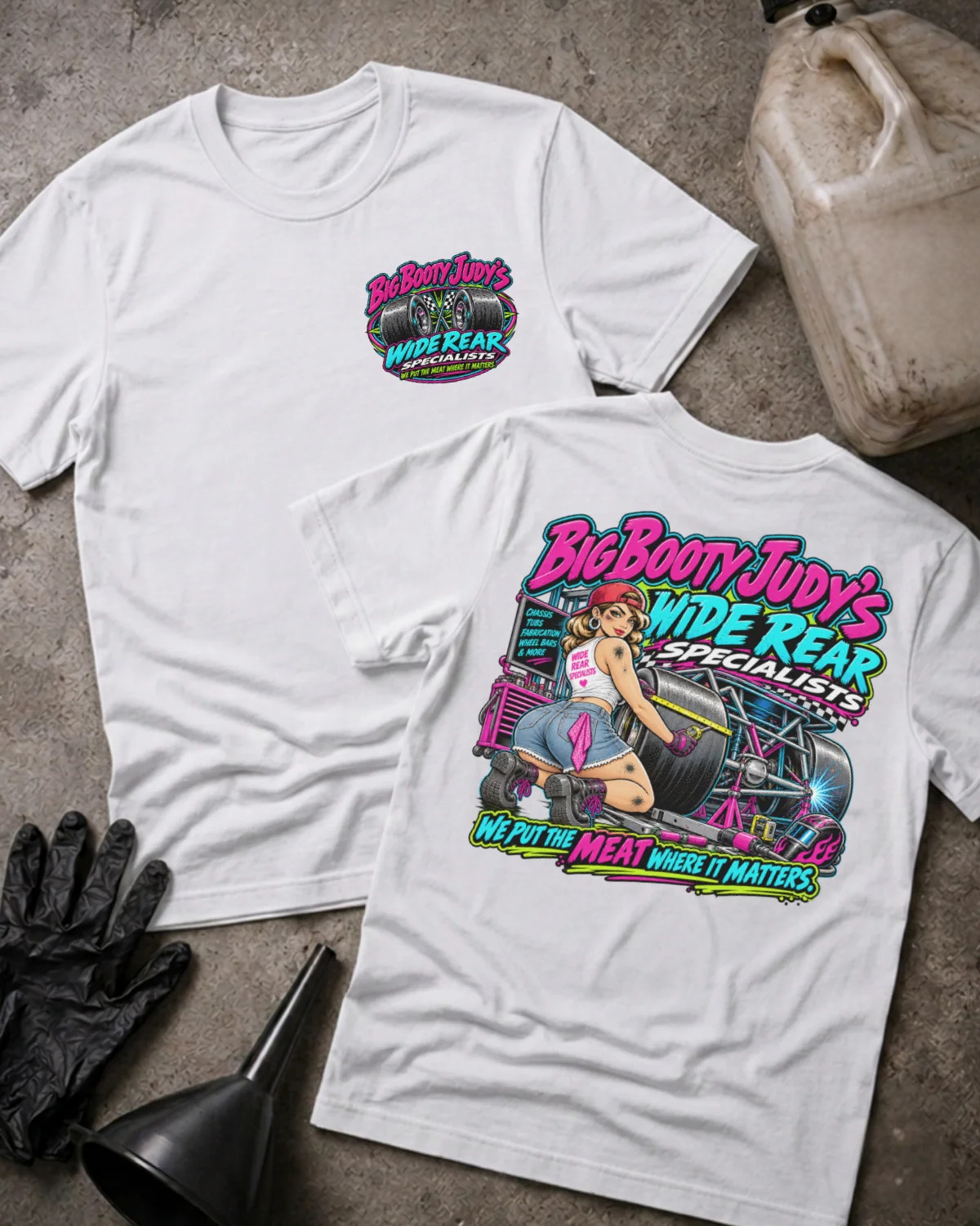 Two white graphic T-shirts laid flat on a concrete surface, showing the front and back designs. The shirts feature a colorful 'Big Booty Judy's Wide Rear Specialist' graphic with a pin-up illustration and hot rod elements.