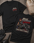 Two black graphic T-shirts laid flat on a textured gray surface with automotive props. The front shirt shows a large graphic of a woman seated on a race car with the text 'Big Chassis Cassie' and 'Wider Where It Counts.
