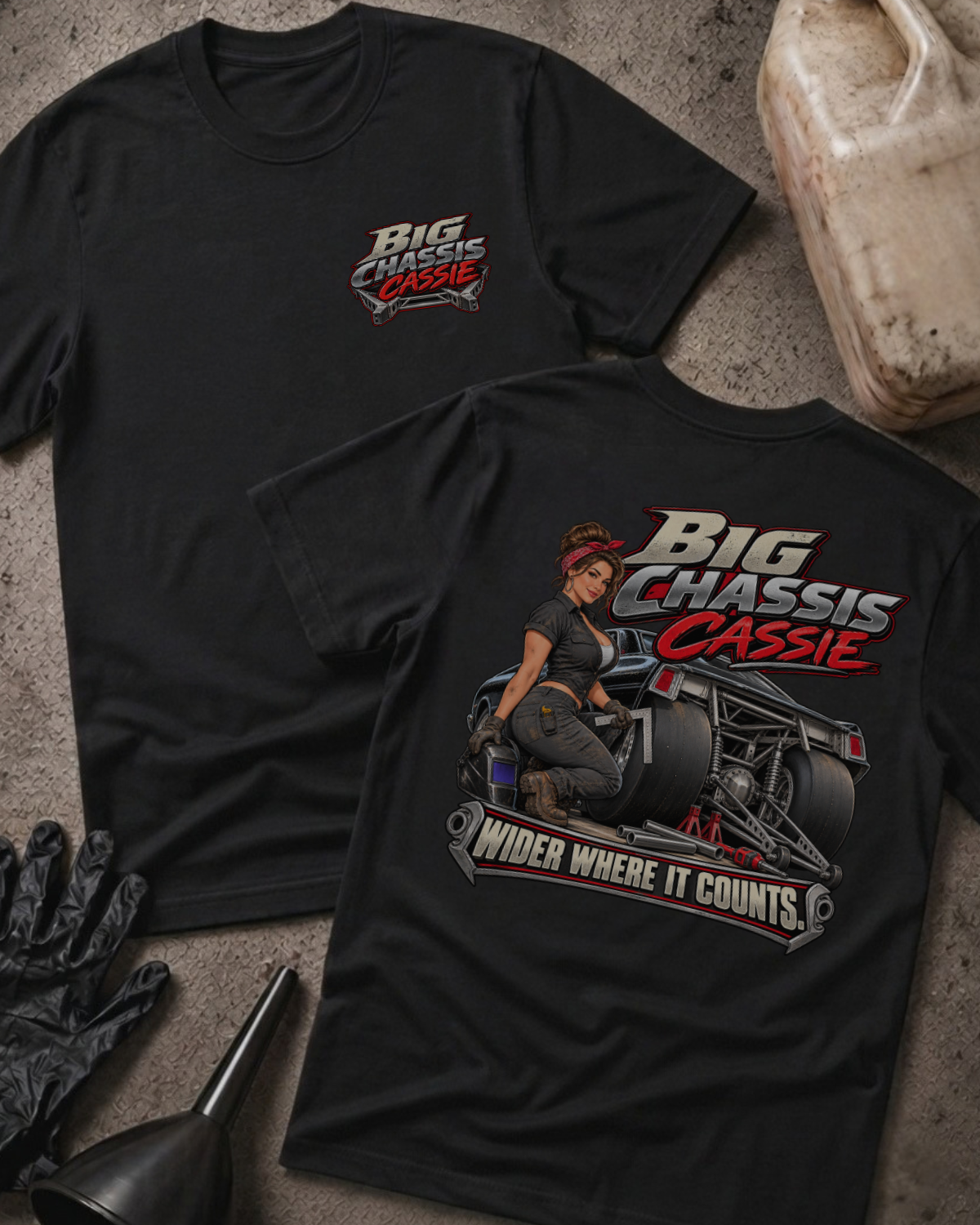 Two black graphic T-shirts laid flat on a textured gray surface with automotive props. The front shirt shows a large graphic of a woman seated on a race car with the text 'Big Chassis Cassie' and 'Wider Where It Counts.