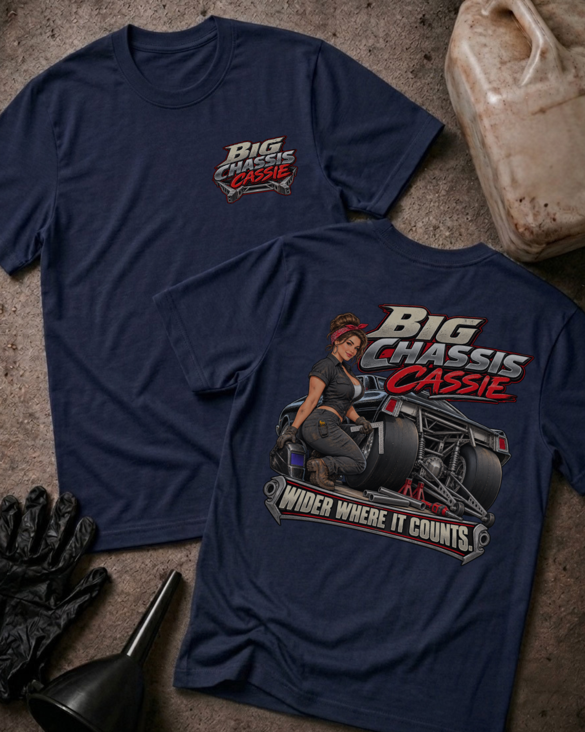 Two navy graphic T-shirts laid flat on a textured gray surface with automotive props. The front shirt shows a large graphic of a woman seated on a race car with the text 'Big Chassis Cassie' and 'Wider Where It Counts.