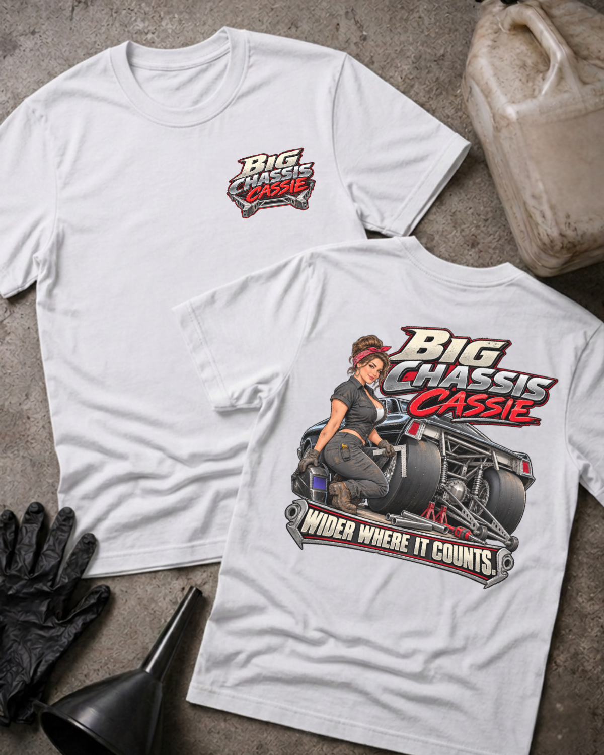 Two white graphic T-shirts laid flat on a textured gray surface with automotive props. The front shirt shows a large graphic of a woman seated on a race car with the text 'Big Chassis Cassie' and 'Wider Where It Counts.