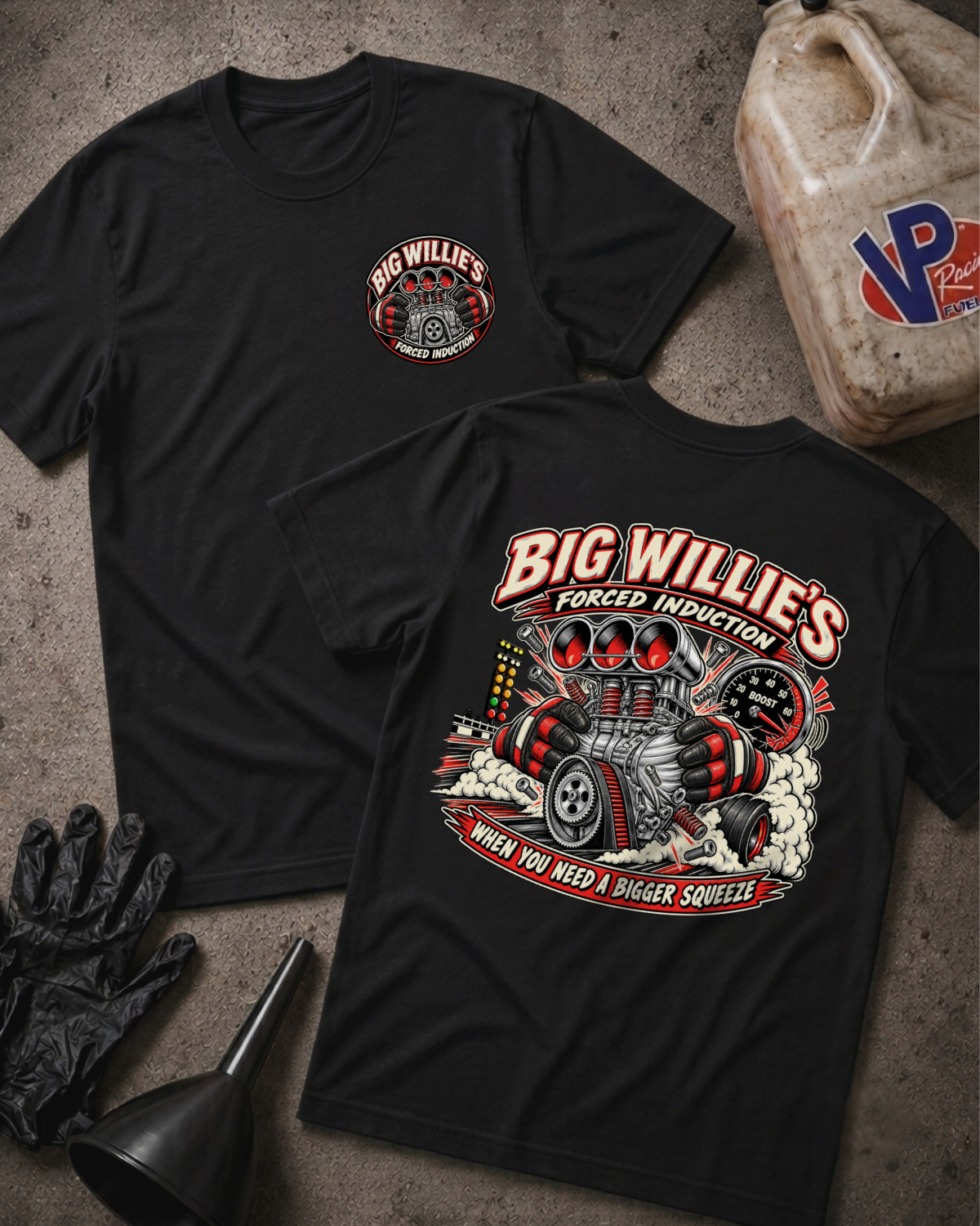 Two black graphic T-shirts laid flat on a textured gray surface, with automotive-themed artwork and the text 'Big Willie's Forced Induction' on the front and back. The scene includes styling props such as a plastic jug, black gloves, and a funnel.