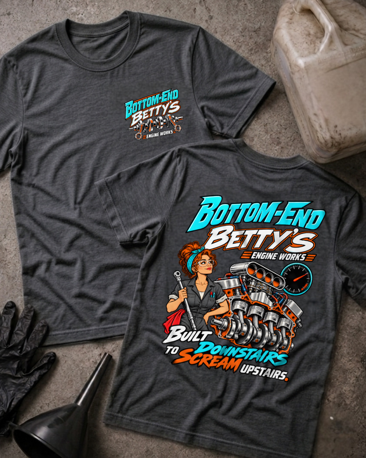 Dark grey heather Bottom End Betty’s Engine Works T-shirt showing the front chest logo and back cartoon of a woman mechanic with a torque wrench by a V8 engine’s crankshaft and pistons, “Built Downstairs to Scream Upstairs,” with gloves, funnel, and oil jug.