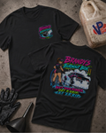 Two black graphic t-shirts displayed on a textured concrete surface with automotive-themed props like a jerrycan, gloves, and funnel. The front shirt features a large colorful graphic of a woman driving a race car with the text 'Brandy's Burnout Box' and 'Wet Your Rubbers.