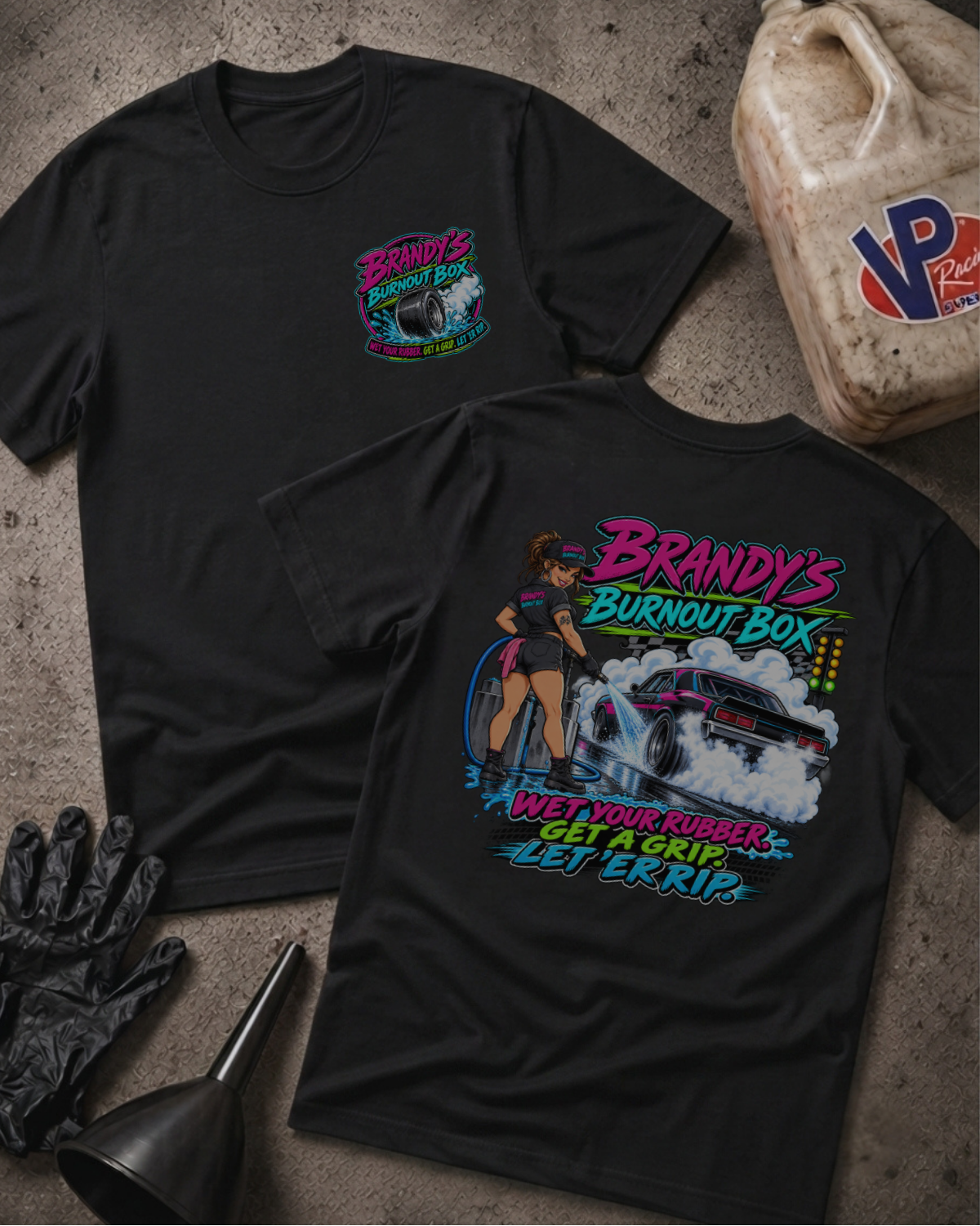 Two black graphic t-shirts displayed on a textured concrete surface with automotive-themed props like a jerrycan, gloves, and funnel. The front shirt features a large colorful graphic of a woman driving a race car with the text 'Brandy's Burnout Box' and 'Wet Your Rubbers.