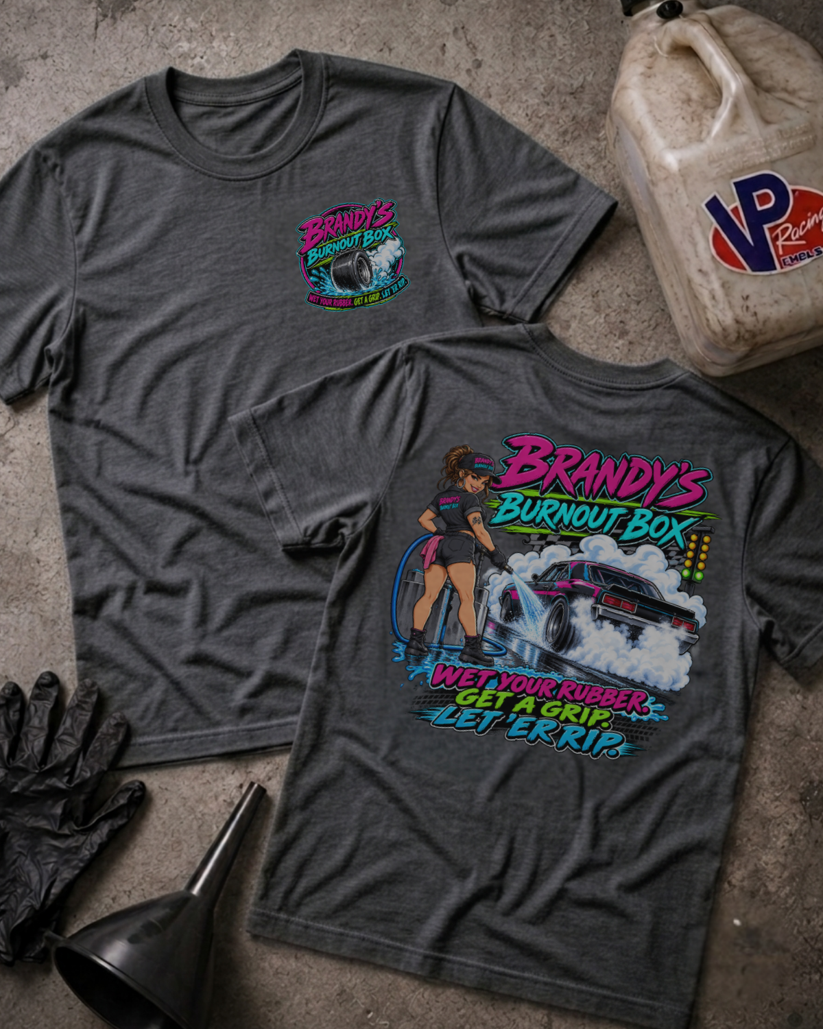 Two dark gray graphic T-shirts laid flat on a concrete surface, with a vintage oil can, black gloves, and a funnel around them. The front shirt shows a small multicolor 'Brandy's Burnout Box' chest graphic, and the back shirt shows a large colorful graphic of a woman standing next to a car with smoke and the text 'Brandy's Burnout Box Wet Your Rubber Get a Grip Let 'Er Rip!'.