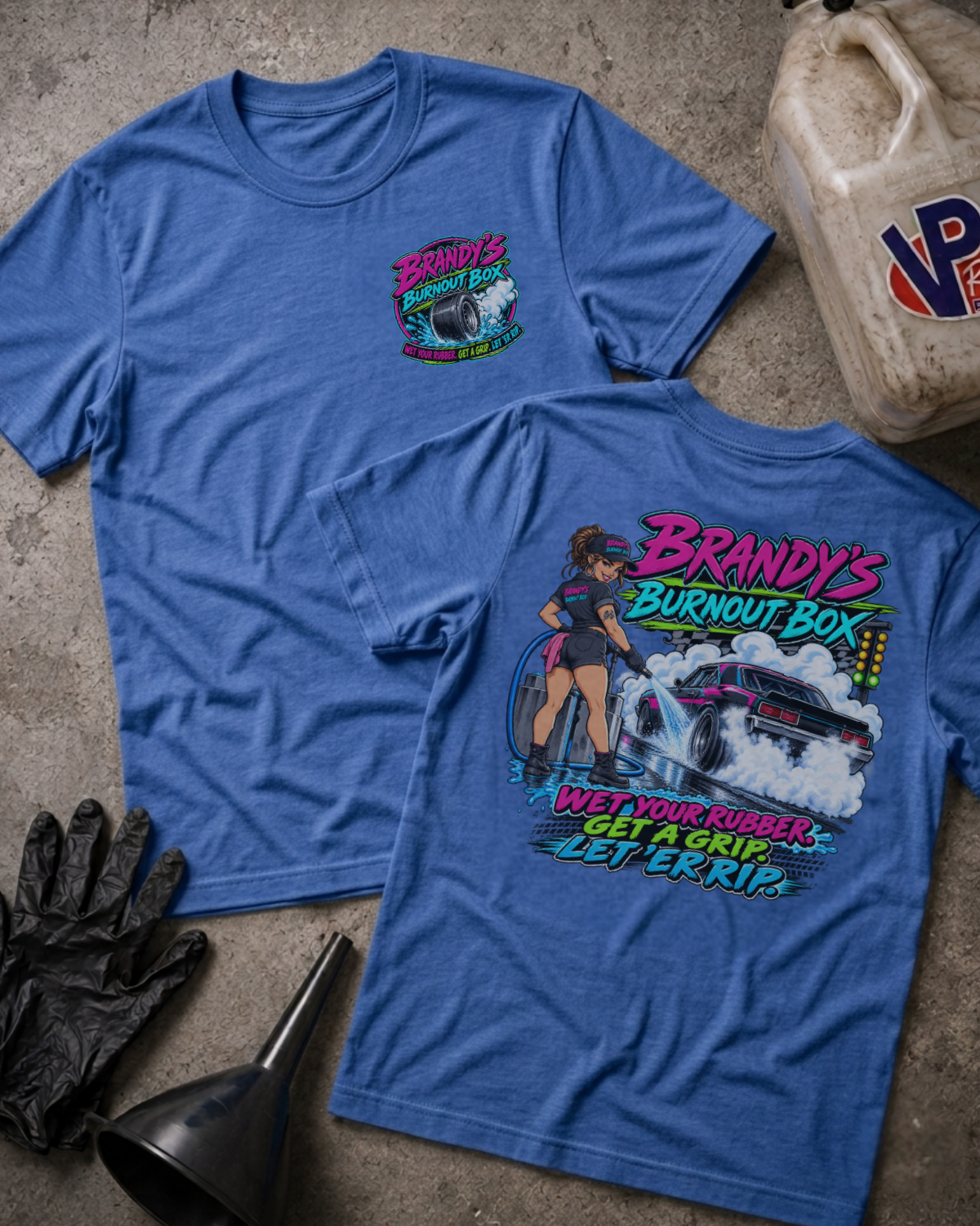 Two heather blue graphic t-shirts laid flat on a concrete surface, showing the front and back designs. The front shirt features a small colorful chest graphic, while the back shirt features a large graphic of a woman driving a race car with the text 'Brandy's Burnout Box' and 'Wet Your Rubber, Get A Grip, Let 'Er Rip.