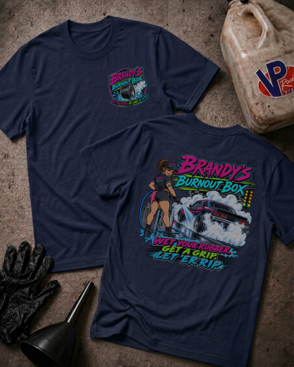 Two navy graphic t-shirts laid flat on a concrete surface, showing the front and back designs. The front shirt features a small colorful chest graphic, while the back shirt features a large graphic of a woman driving a race car with the text 'Brandy's Burnout Box' and 'Wet Your Rubber, Get A Grip, Let 'Er Rip.