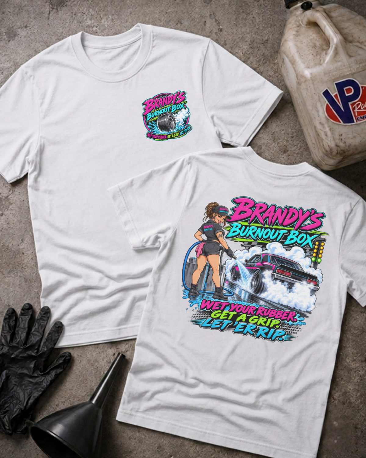 Two white graphic T-shirts displayed on a concrete floor with automotive-themed props, showing both the front and back designs. The front features a small colorful chest graphic, and the back features a large graphic with the text 'Brandy's Burnout Box' and a race car illustration.