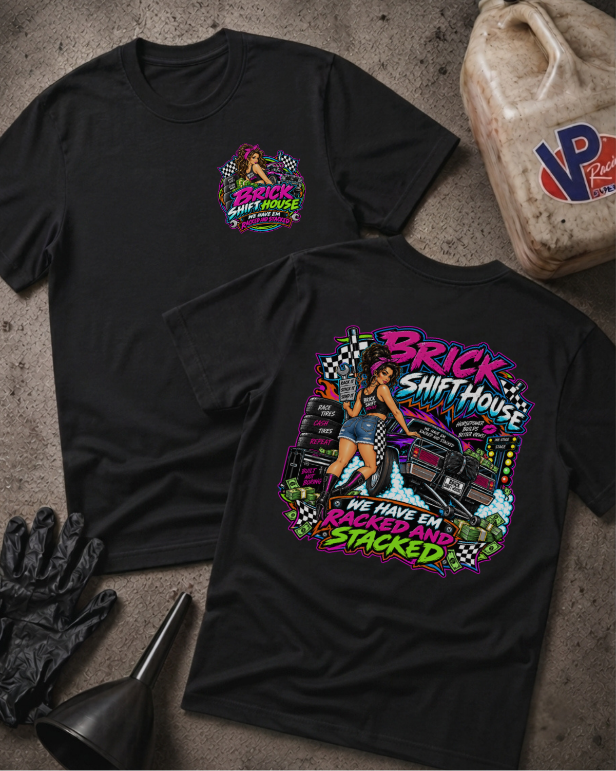 Two black graphic T-shirts laid flat on a textured concrete surface, with a gas bottle, black gloves, and funnel as styling props. The front shirt shows a small multicolor chest graphic, and the back shirt shows a large colorful graphic with the words 'Brick Shift House' and 'We Have Em Racked And Stacked'.