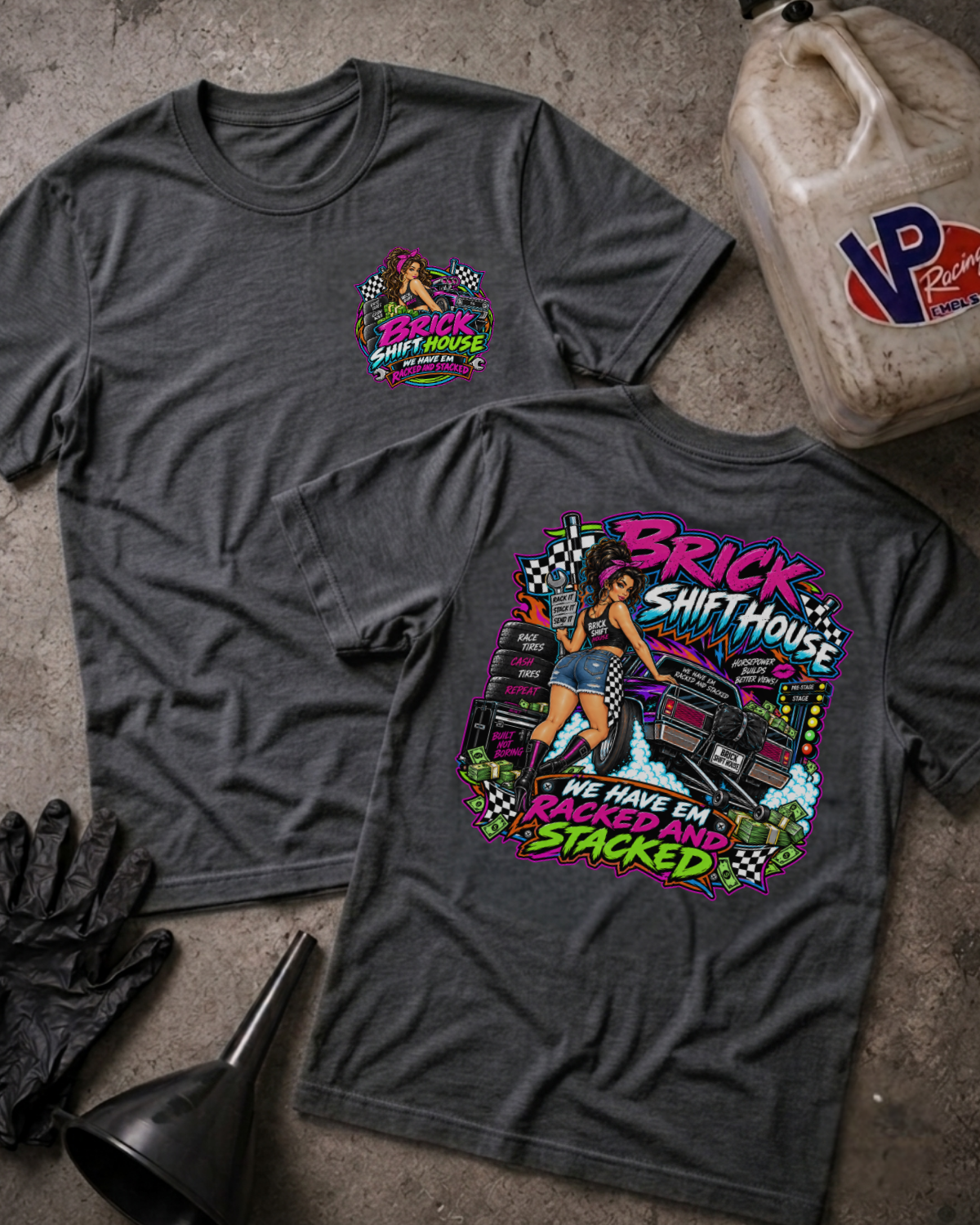 Two dark grey heather graphic T-shirts laid flat on a textured concrete surface, with a gas bottle, black gloves, and funnel as styling props. The front shirt shows a small multicolor chest graphic, and the back shirt shows a large colorful graphic with the words 'Brick Shift House' and 'We Have Em Racked And Stacked'.