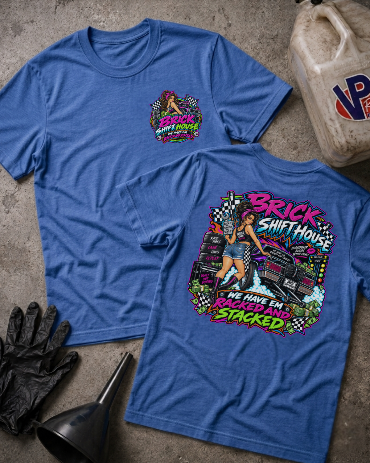 Two blue graphic T-shirts laid flat on a concrete-like surface, with a black glove, funnel, and a worn plastic jug nearby. The front shirt shows a small multicolor chest graphic, and the back shirt shows a large colorful graphic with the words 'Brick Shift House' and 'We Have Em Racked and Stacked'