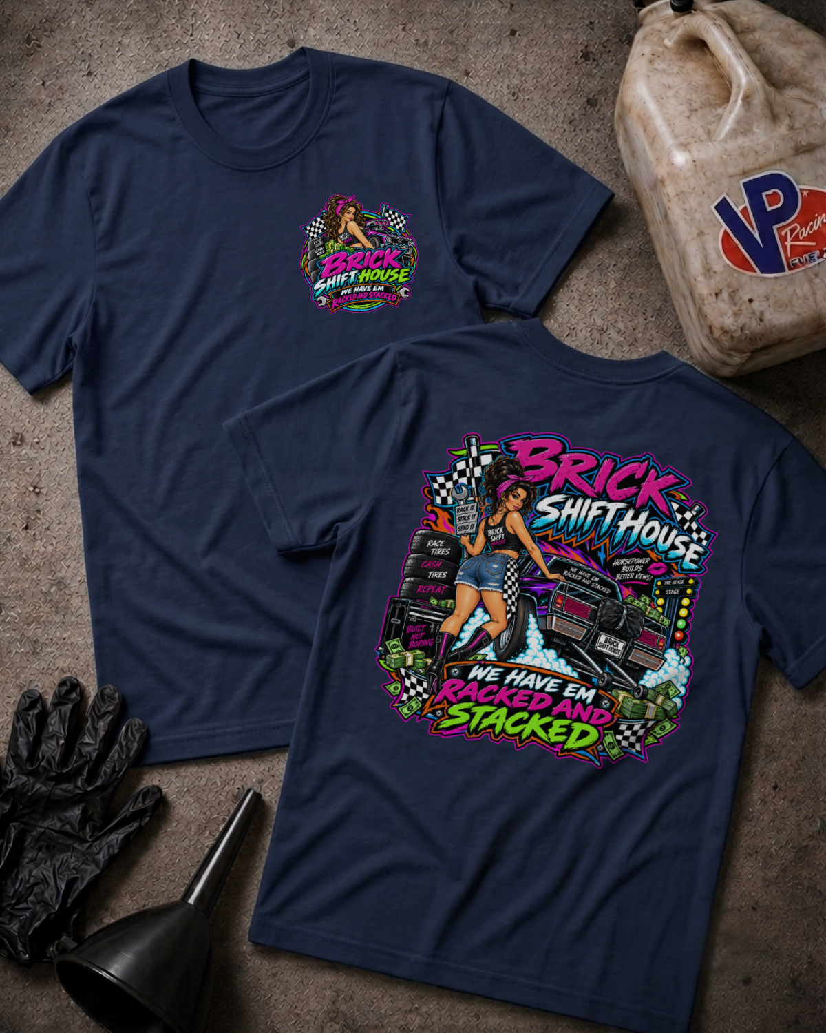Two navy blue graphic t-shirts displayed on a textured concrete floor with racing-themed props, including a jerrycan, gloves, and funnel. The front shirt shows a small chest graphic, while the back shirt features a large colorful graphic with the text 'BRICK SHIFT HOUSE' and 'WE HAVE EM RACKED AND STACKED'.