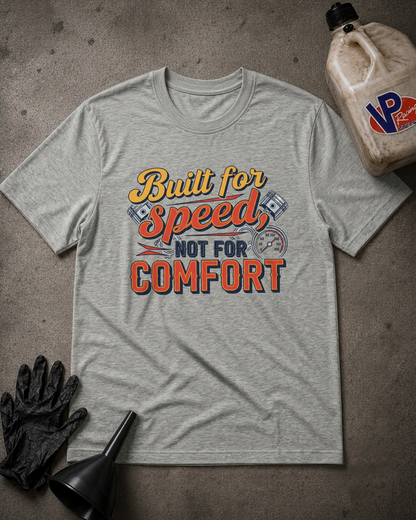 A heather gray graphic t-shirt laid flat on a textured gray surface, featuring the text 'BUILT FOR SPEED, NOT FOR COMFORT' in yellow, blue, and red. The shirt is styled with a black glove, a black funnel, and a jug of VP Performance oil in the corners.
