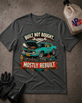 A dark heather gray graphic t-shirt laid flat on a textured gray surface, featuring a teal muscle car illustration with the text 'BUILT NOT BOUGHT.