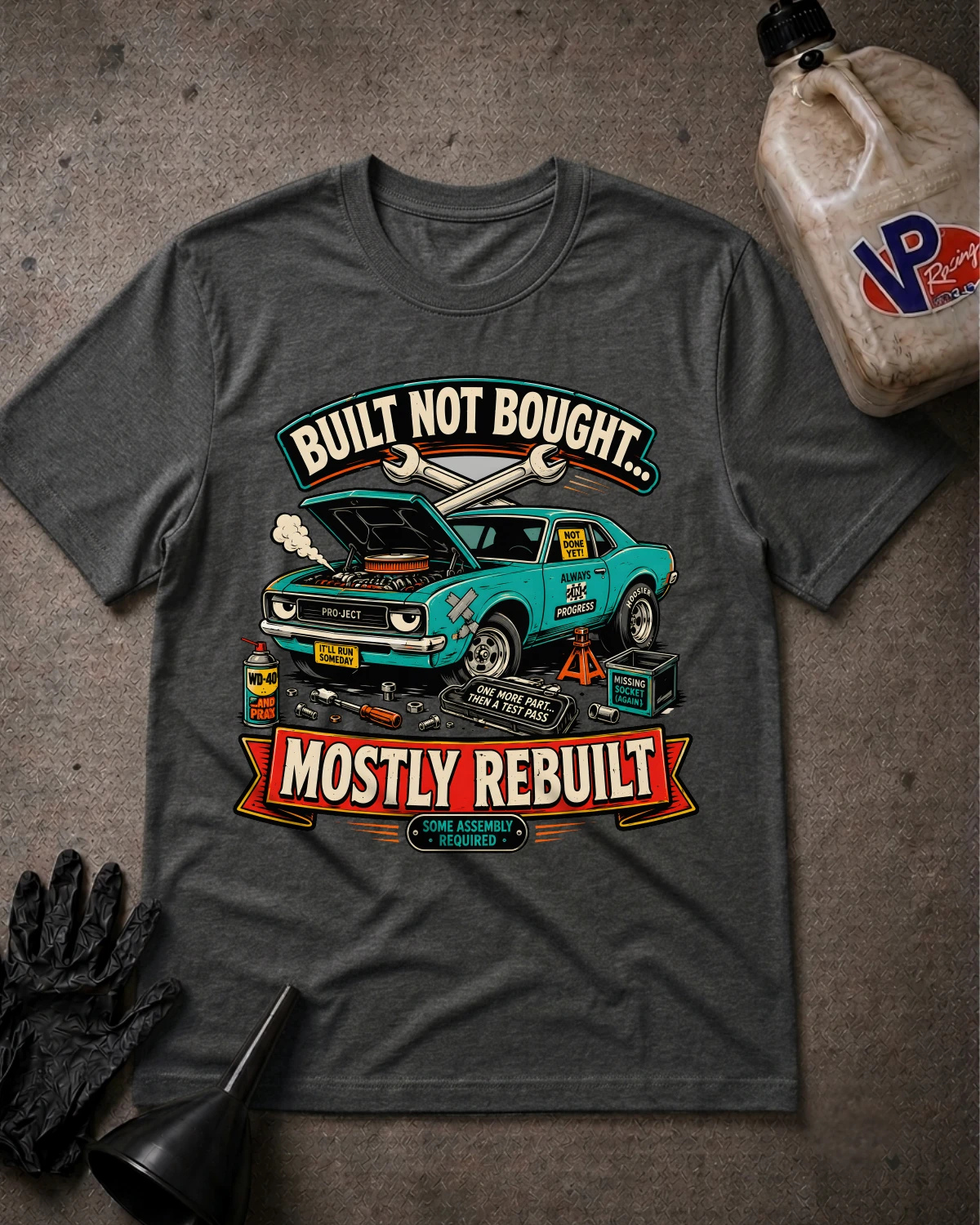 A dark heather gray graphic t-shirt laid flat on a textured gray surface, featuring a teal muscle car illustration with the text 'BUILT NOT BOUGHT.