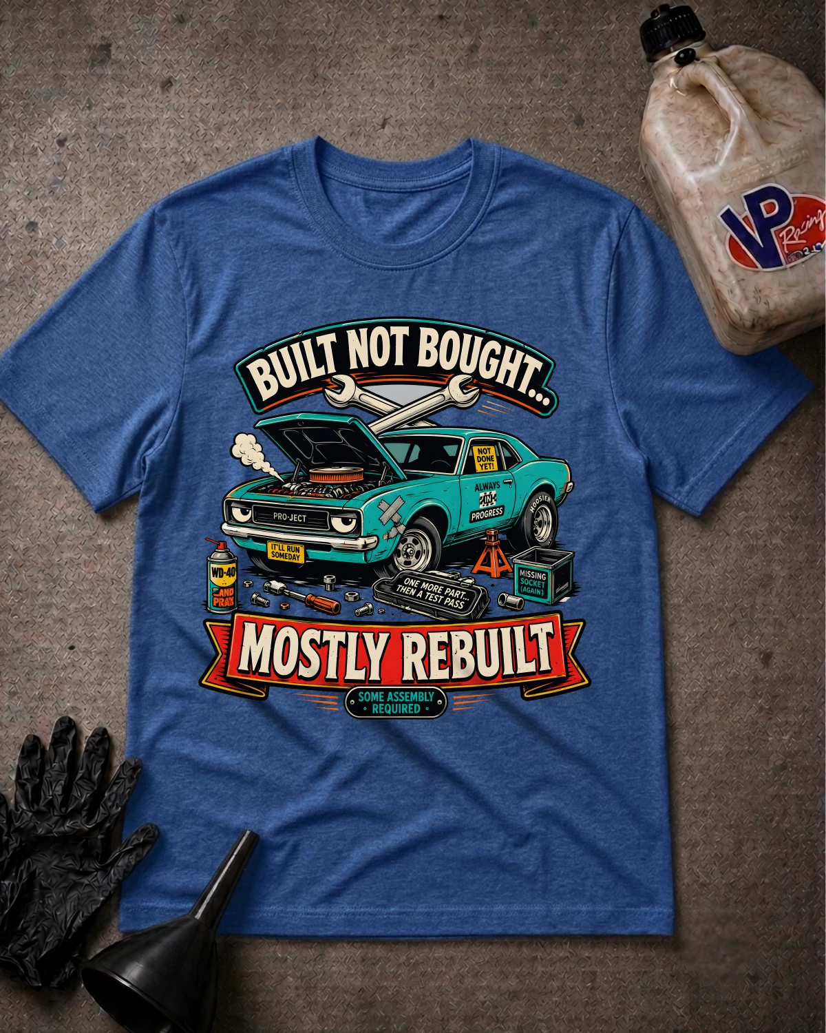 A blue graphic t-shirt laid flat on a textured gray surface, featuring a teal car illustration with the text 'BUILT NOT BOUGHT.