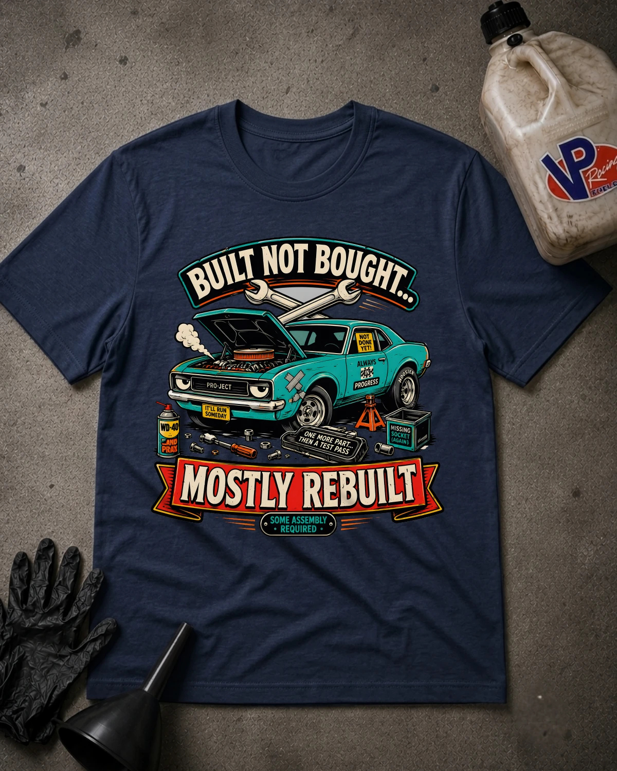 A navy blue graphic t-shirt laid flat on a textured gray surface, surrounded by automotive-themed props including a black oil pan, black gloves, and a plastic jug. The t-shirt features a graphic of a turquoise muscle car with its hood open, with the text 'BUILT NOT BOUGHT.