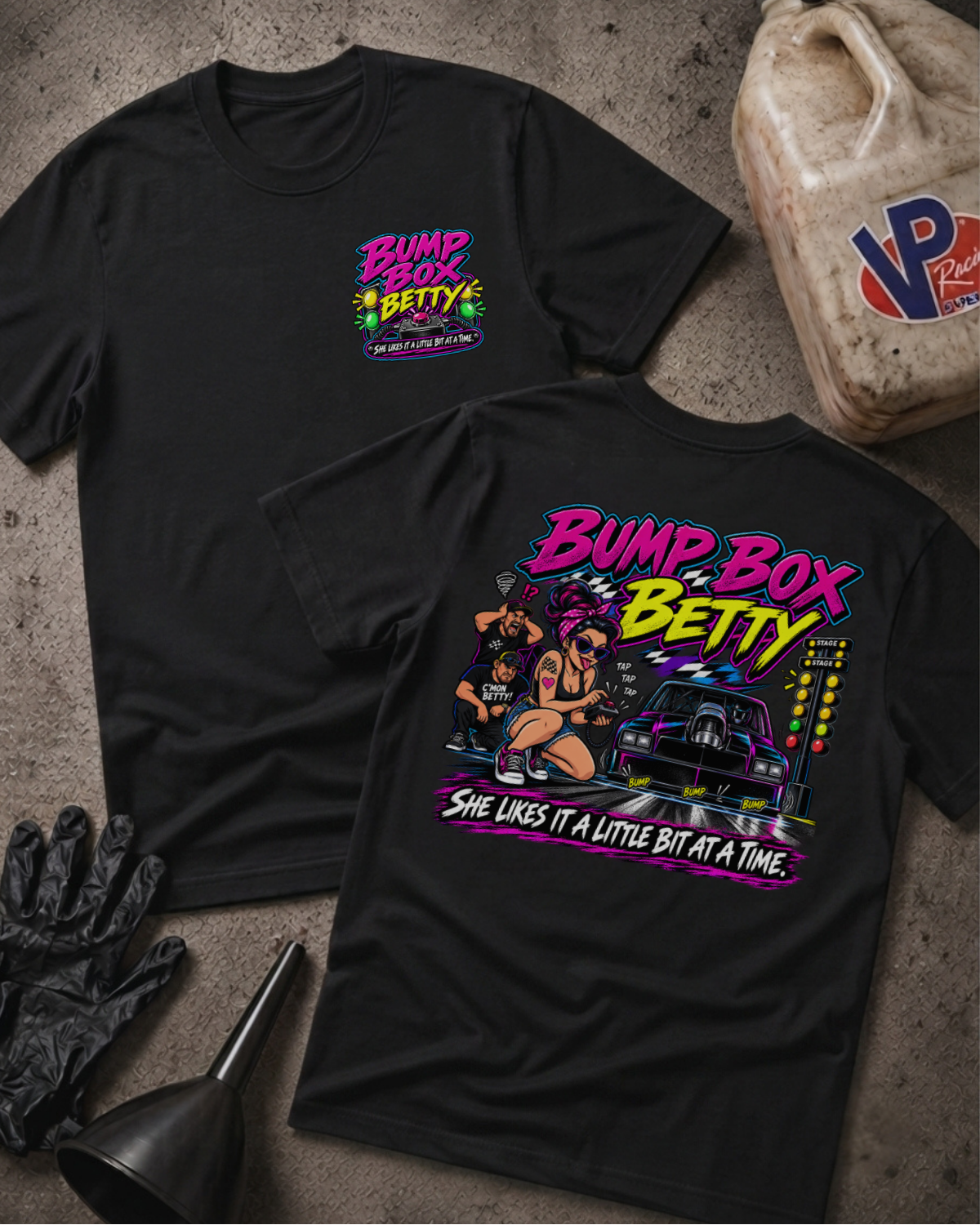 Two black graphic T-shirts laid flat on a textured concrete surface, with a gas can, black gloves, and a funnel as styled props. The front shirt shows a small colorful chest print reading 'Bump Box Betty' with 'She Likes It A Little Bit At A Time' below, and the back shirt shows a large multicolor graphic with the same text and an illustrated woman beside a car.