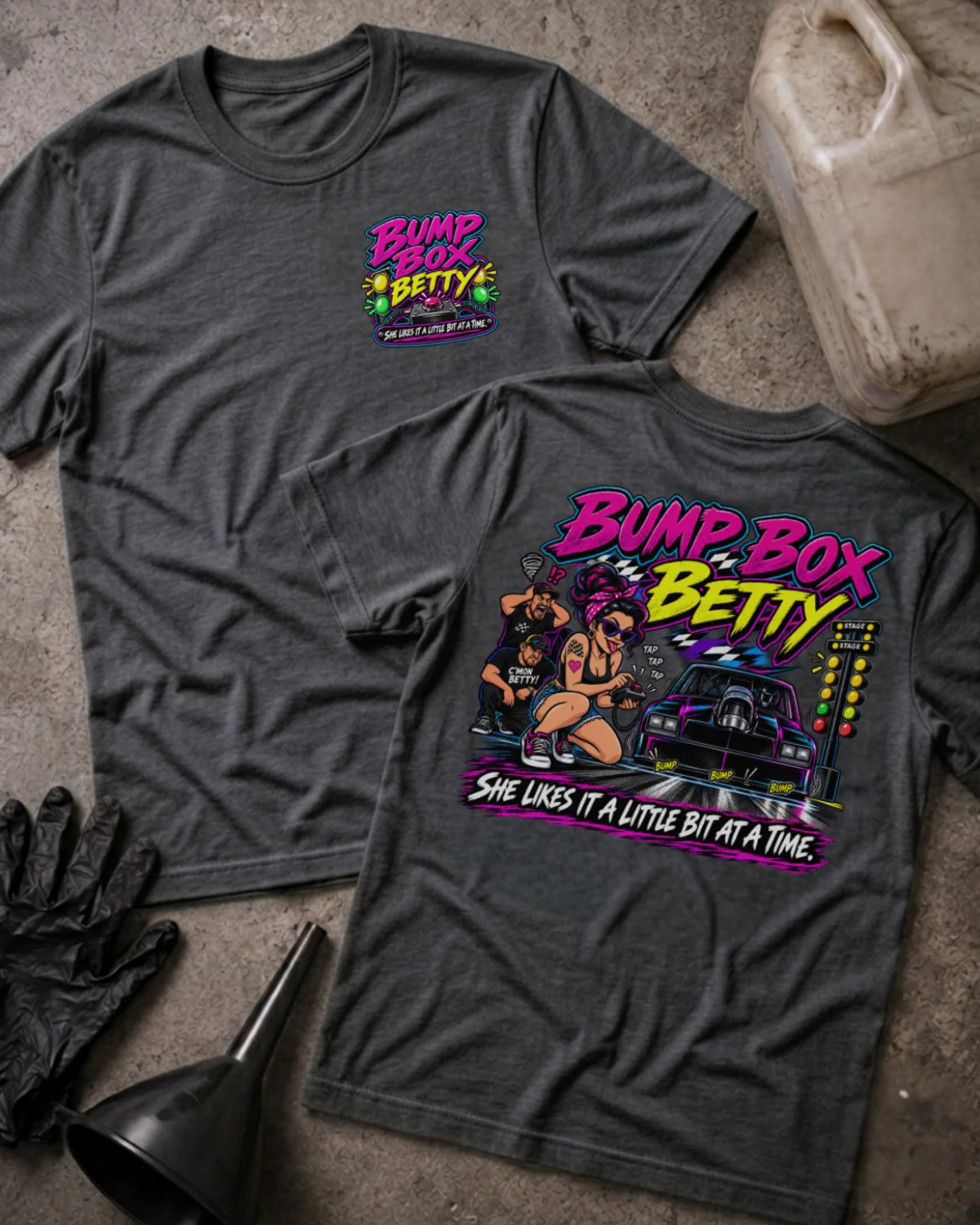 Front and back of Bump Box Betty dark grey heather T-shirt with chest logo, woman using a handheld controller beside a drag car and staging lights, and “She likes it a little bit at a time” slogan.