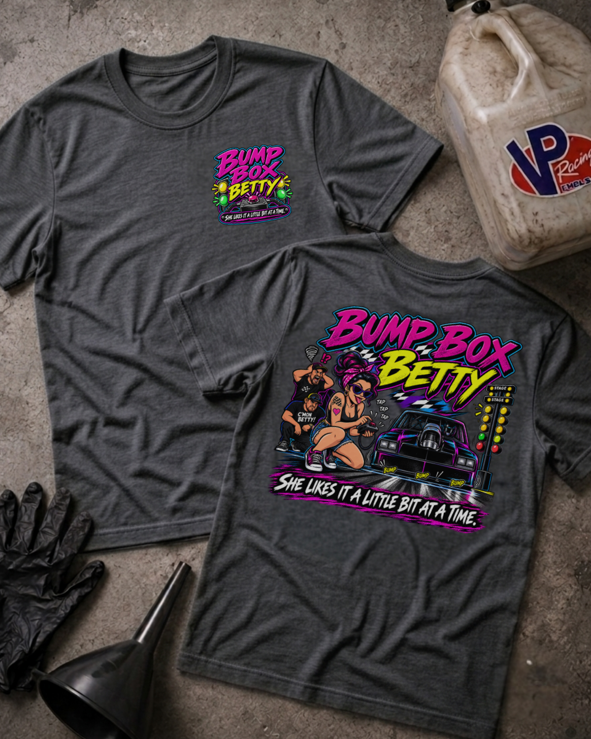 Two dark heather gray graphic T-shirts laid flat on a concrete surface, showing the front and back designs. The shirts feature a colorful 'Bump Box Betty' graphic with a car, illustrated figures, and the phrase 'She likes it a little bit at a time.