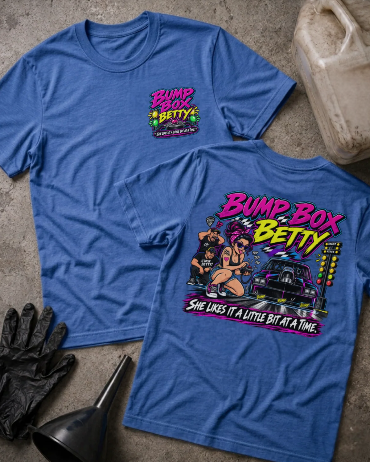 Front and back of Bump Box Betty heather true royal T-shirt with chest logo, woman using a handheld controller beside a drag car and staging lights, and “She likes it a little bit at a time” slogan.