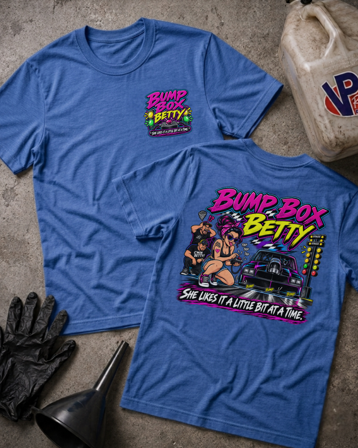 Two heather blue graphic T-shirts laid flat on a concrete surface, showing both front and back designs. The front has a small multicolor chest graphic reading 'Bump Box Betty,' and the back has a large colorful graphic with the same text plus an illustrated character, car, and smaller text.