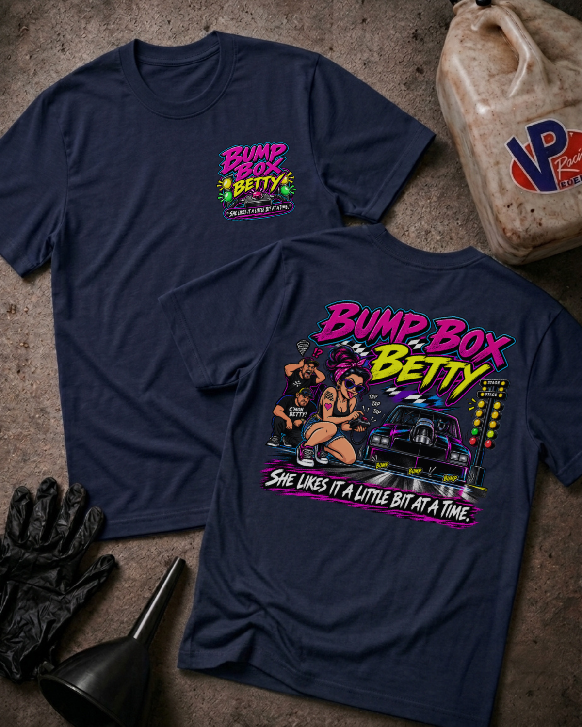 Two navy graphic T-shirts laid flat on a concrete surface, showing the front and back designs. The shirts feature a colorful 'Bump Box Betty' graphic with a car, illustrated figures, and the phrase 'She likes it a little bit at a time.