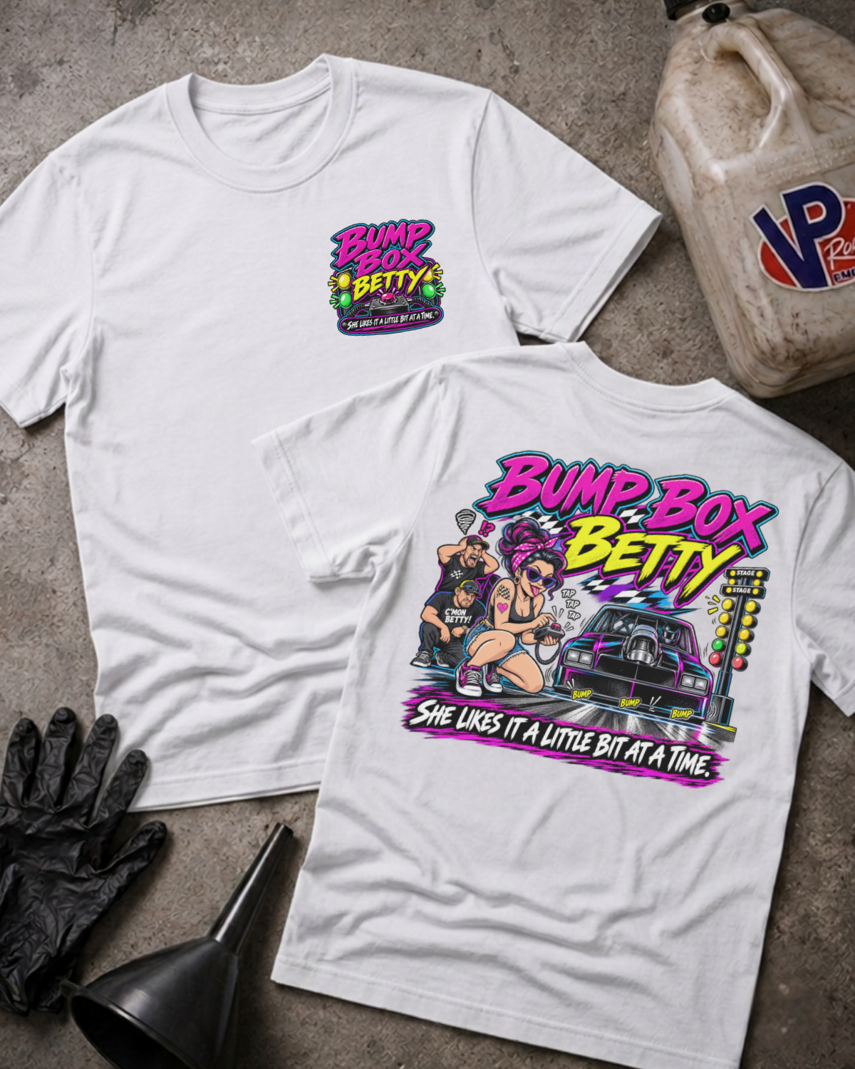 Two white graphic T-shirts laid flat on a concrete surface, showing the front and back designs. The front has a small colorful 'Bump Box Betty' chest graphic, and the back has a large multicolor graphic with the same text plus an illustrated hot rod scene.
