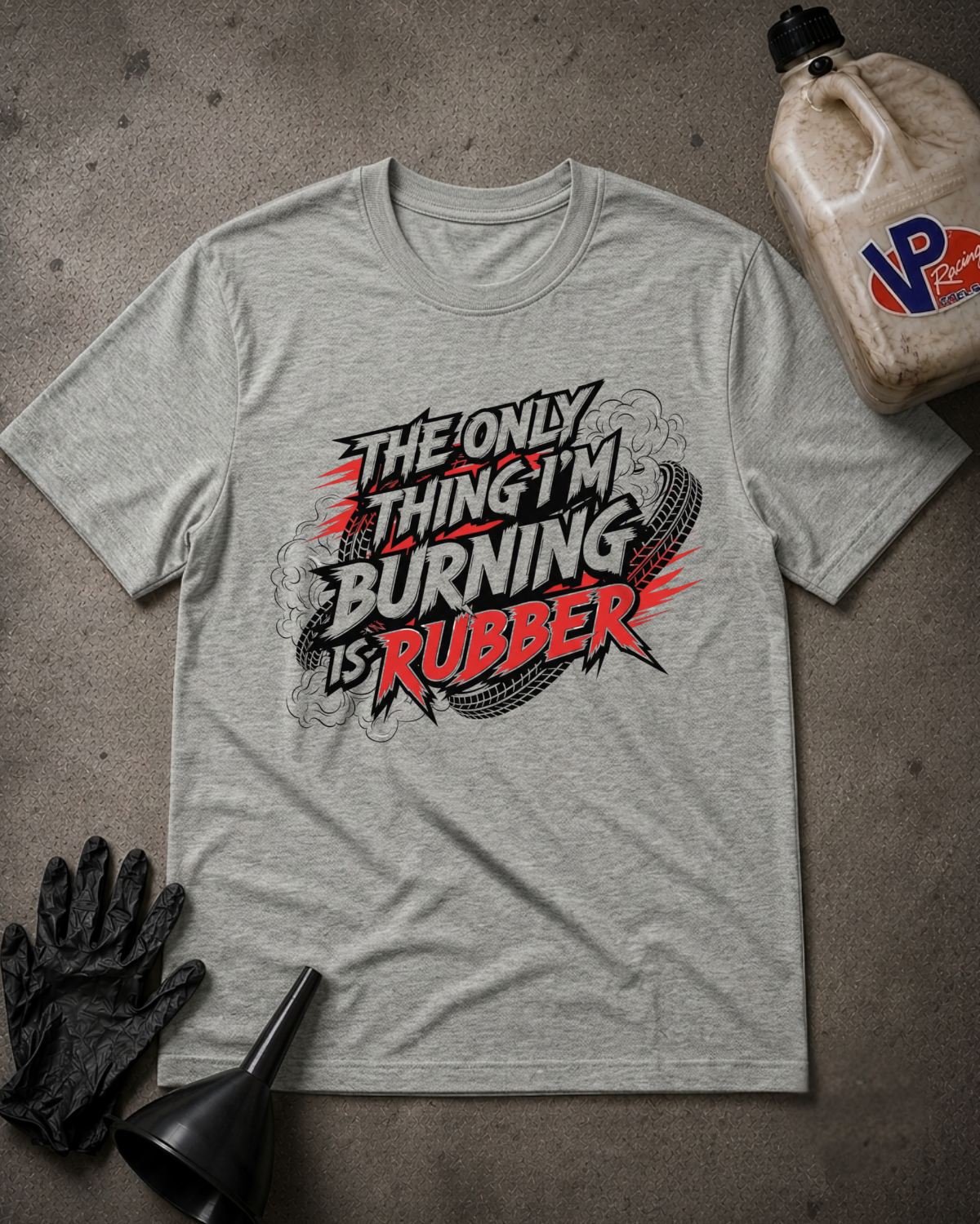 Athletic heather graphic t-shirt laid flat on a dark textured surface, surrounded by race-themed props including a black oil filter wrench, a black work glove, and a jug of VP Racing oil. The t-shirt features a bold graphic reading 'THE ONLY THING I'M BURNING IS RUBBER' in red, black, and white.