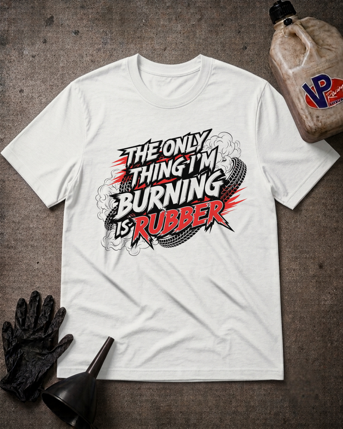 A white graphic t-shirt laid flat on a dark textured surface, surrounded by race-themed props including a black oil filter wrench, a black work glove, and a jug of VP Racing oil. The t-shirt features a bold graphic reading 'THE ONLY THING I'M BURNING IS RUBBER' in red, black, and white.