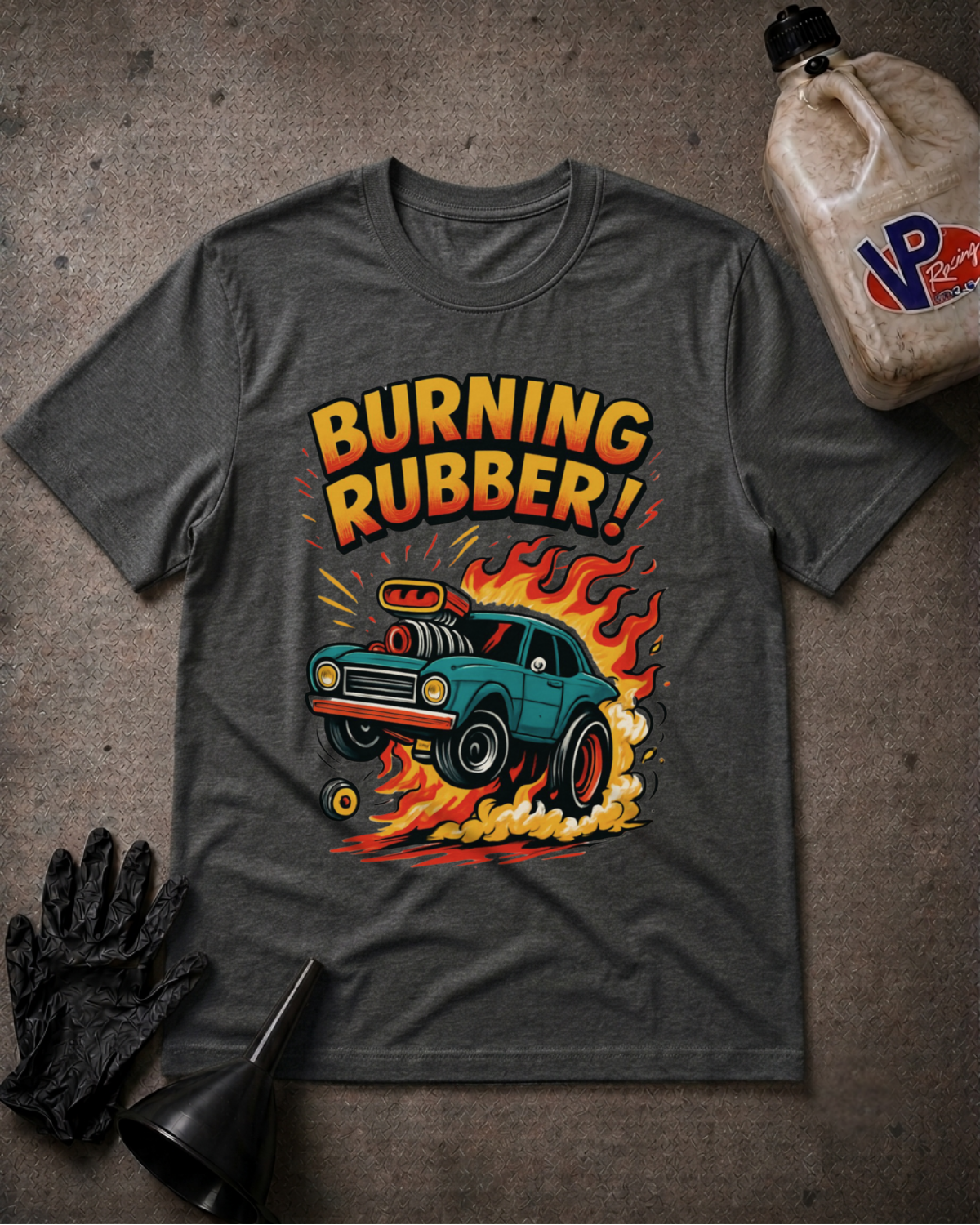 A dark heather gray graphic t-shirt laid flat on a textured gray surface, featuring a retro-style illustration of a teal car with flames and the text 'BURNING RUBBER!'. A black glove, funnel, and a plastic jug labeled 'VP Racing' are arranged around it as styling props.