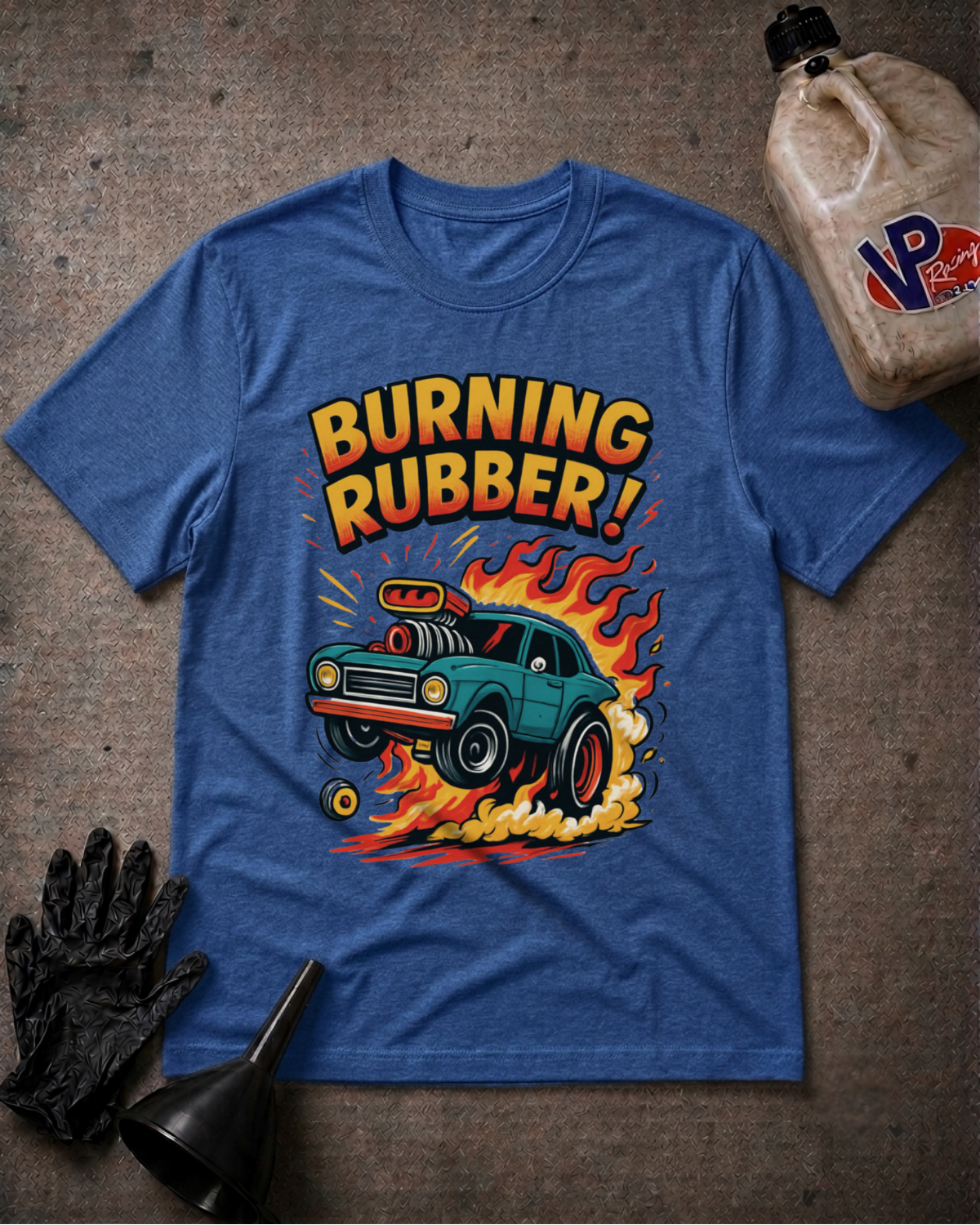 A heather blue graphic t-shirt laid flat on a textured gray surface, featuring a retro-style illustration of a teal car with flames and the text 'BURNING RUBBER!'. A black glove, funnel, and a plastic jug are styled around the shirt.