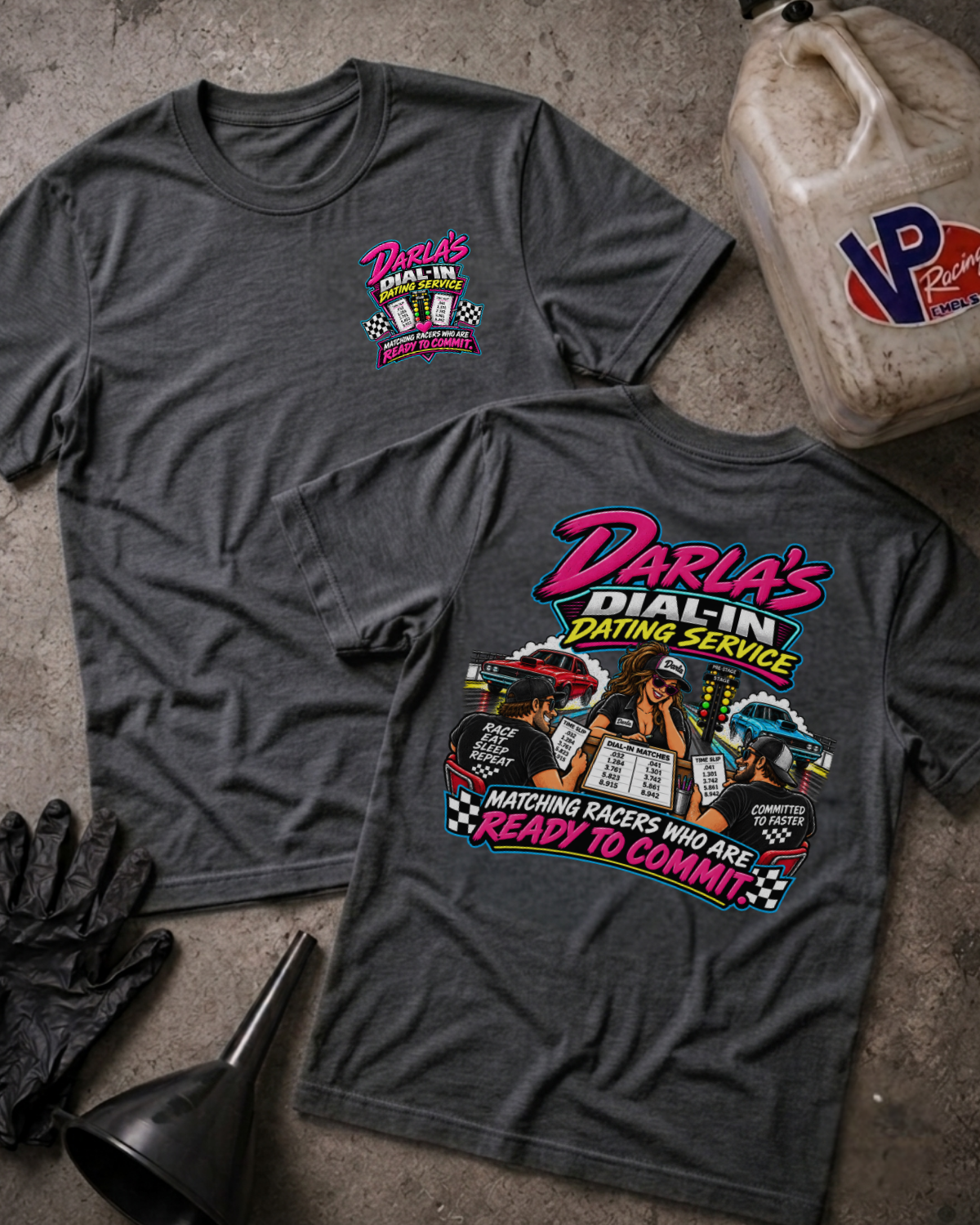 Two dark gray graphic T-shirts laid flat on a concrete surface, with a vintage oil jug, black gloves, and funnel as styled props. The front shirt shows a small pink and yellow chest graphic reading Darla's Dial-In Dating Service, and the back shirt shows a large multicolor racing-themed graphic with the same brand text.