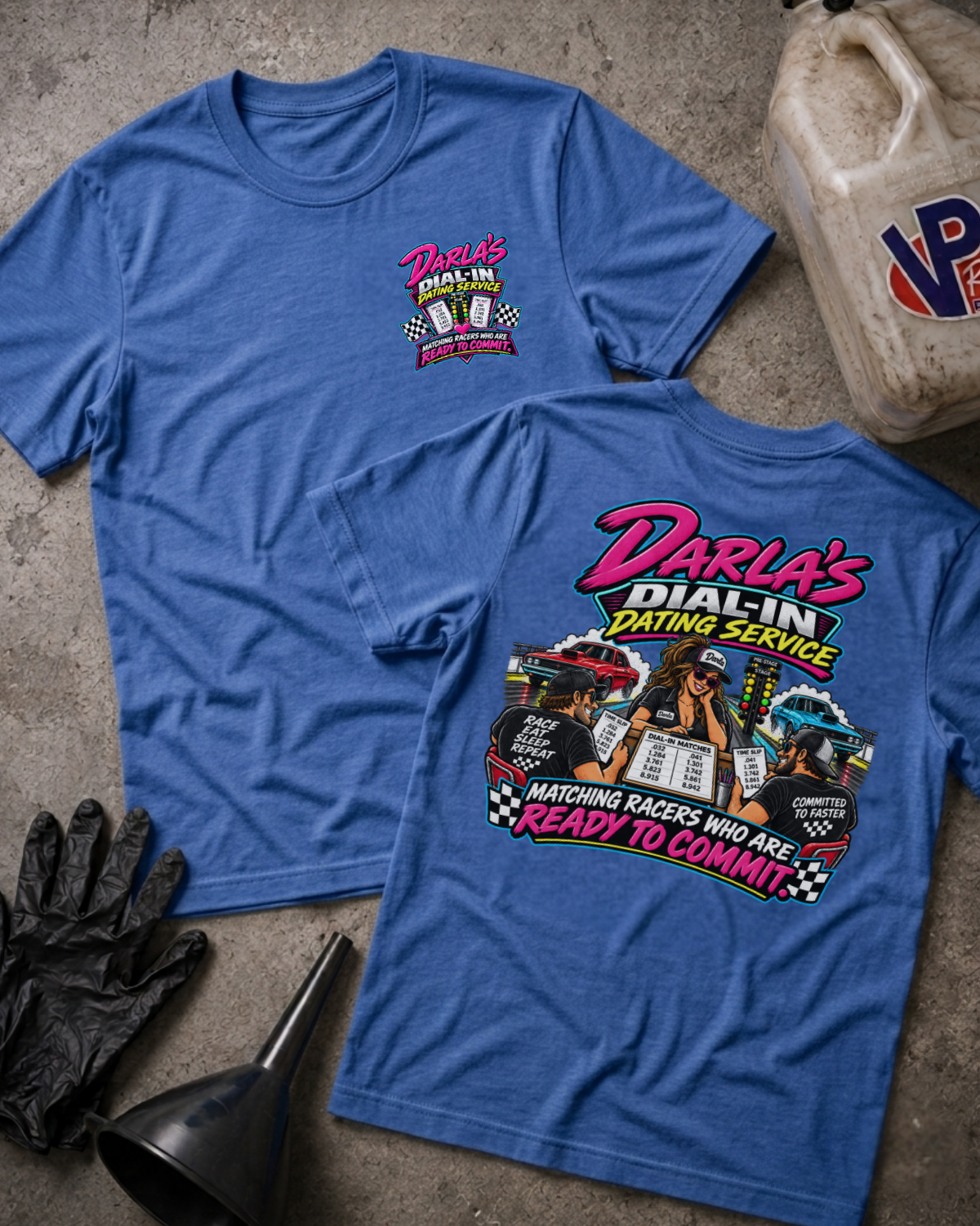 Two heather true royal graphic T-shirts laid flat on a concrete surface, with a vintage oil jug, black gloves, and funnel as styled props. The front shirt shows a small pink and yellow chest graphic reading Darla's Dial-In Dating Service, and the back shirt shows a large multicolor racing-themed graphic with the same brand text.