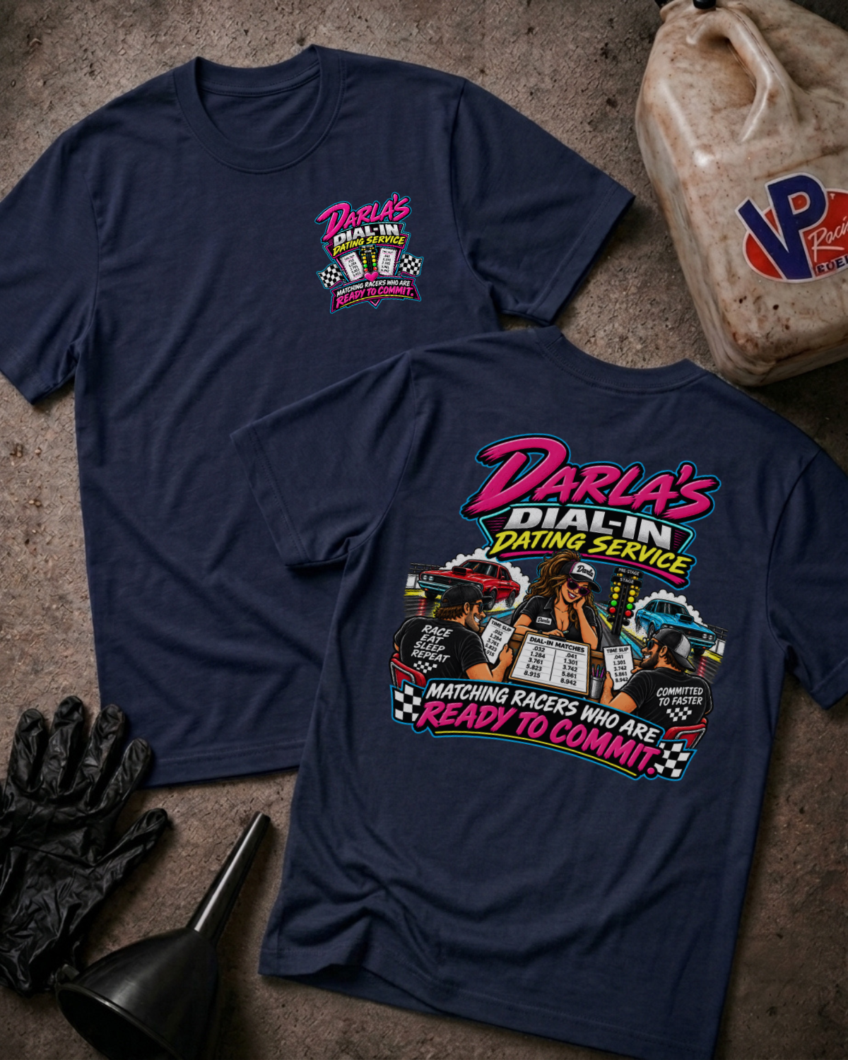 Two navy graphic T-shirts laid flat on a concrete surface, with a vintage oil jug, black gloves, and funnel as styled props. The front shirt shows a small pink and yellow chest graphic reading Darla's Dial-In Dating Service, and the back shirt shows a large multicolor racing-themed graphic with the same brand text.