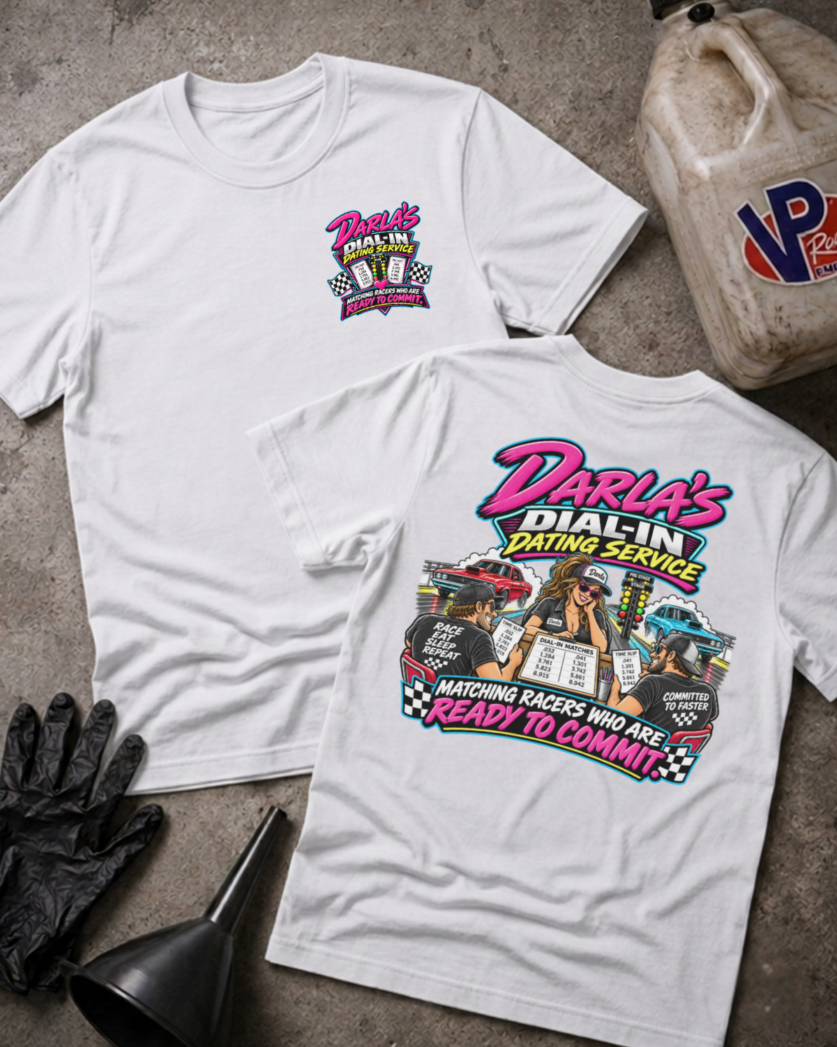 Two white graphic T-shirts laid flat on a concrete surface, with a vintage oil jug, black gloves, and funnel as styled props. The front shirt shows a small pink and yellow chest graphic reading Darla's Dial-In Dating Service, and the back shirt shows a large multicolor racing-themed graphic with the same brand text.
