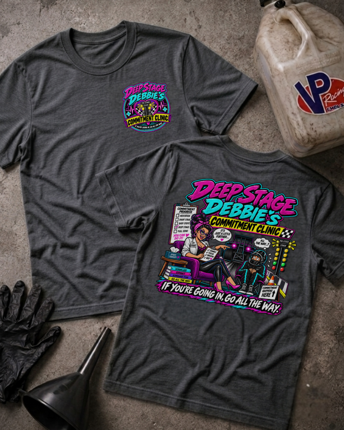 Two dark heather gray graphic T-shirts laid flat on a concrete surface, with a vintage-style oil can, black gloves, and a black funnel as props. The front shirt shows a large colorful comic-style graphic reading 'Deep Stage Debbie's Commitment Clinic' and the slogan 'If you're going in, go all the way!'.
