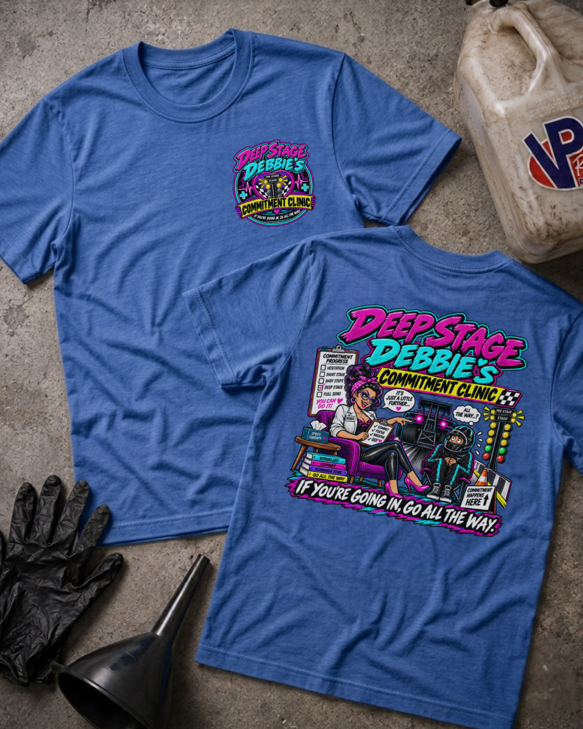 Two heather blue graphic T-shirts laid flat on a concrete surface, showing the front and back designs for 'Deep Stage Debbie's Commitment Center'. The front has a small multicolor chest graphic, and the back has a large colorful comic-style graphic with text.