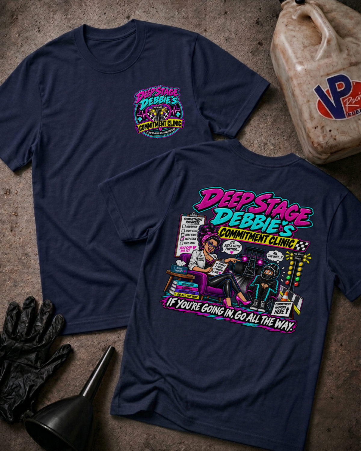Two navy graphic T-shirts displayed on a concrete floor, with a black funnel, black gloves, and a plastic fuel container as props. The front shirt shows a small colorful chest graphic, and the back shirt shows a large colorful graphic with the text 'Deep Stage Debbie's Commitment Clinic' and 'If you're going in, go all the way.