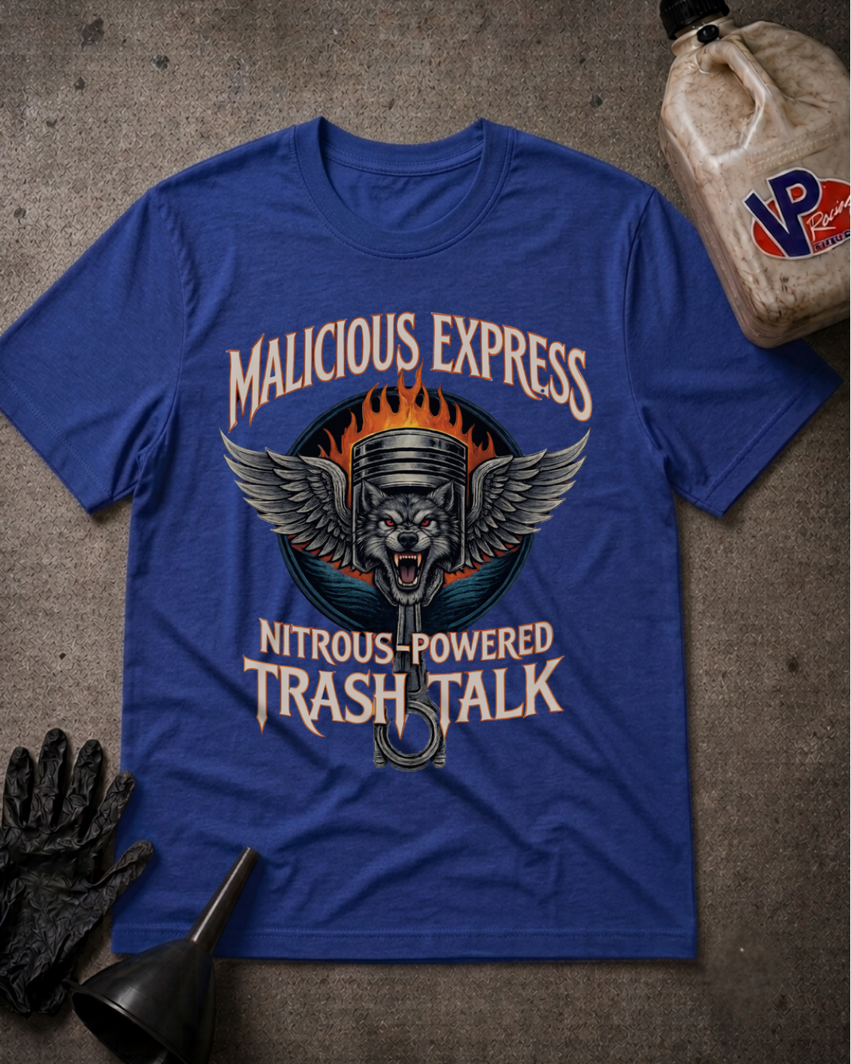 A royal blue graphic t-shirt laid flat on a textured gray concrete surface, surrounded by racing-themed props including an oil bottle, a black glove, a funnel. The shirt features a graphic of a wolf with wings and a piston, with the text 'MALICIOUS EXPRESS' and 'NITROUS-POWERED TRASH TALK'.