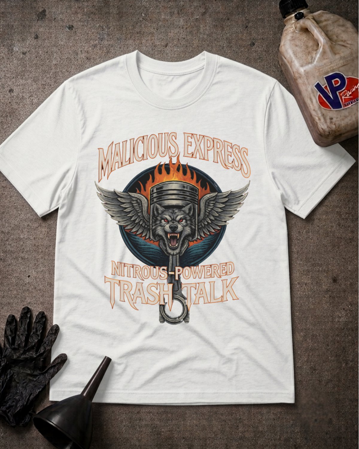 A white graphic t-shirt laid flat on a dark textured surface, surrounded by motorcycle-themed props including an a black funnel, a black glove, and a plastic jug. The t-shirt features a central graphic of a winged piston with flames and the text 'MALICIOUS EXPRESS' and 'NITROUS POWERED TRASH TALK'.