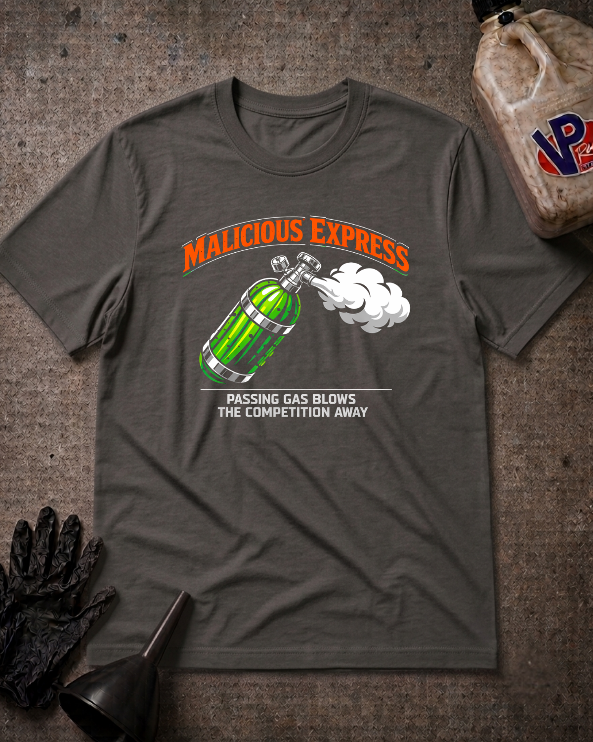 An asphalt graphic t-shirt laid flat on a textured gray surface, featuring a green nitrous bottle illustration with white smoke and the text 'MALICIOUS EXPRESS' and 'PASSING GAS BLOWS THE COMPETITION AWAY'. A gas bottle, funnel, and black glove are styled as props around the shirt.