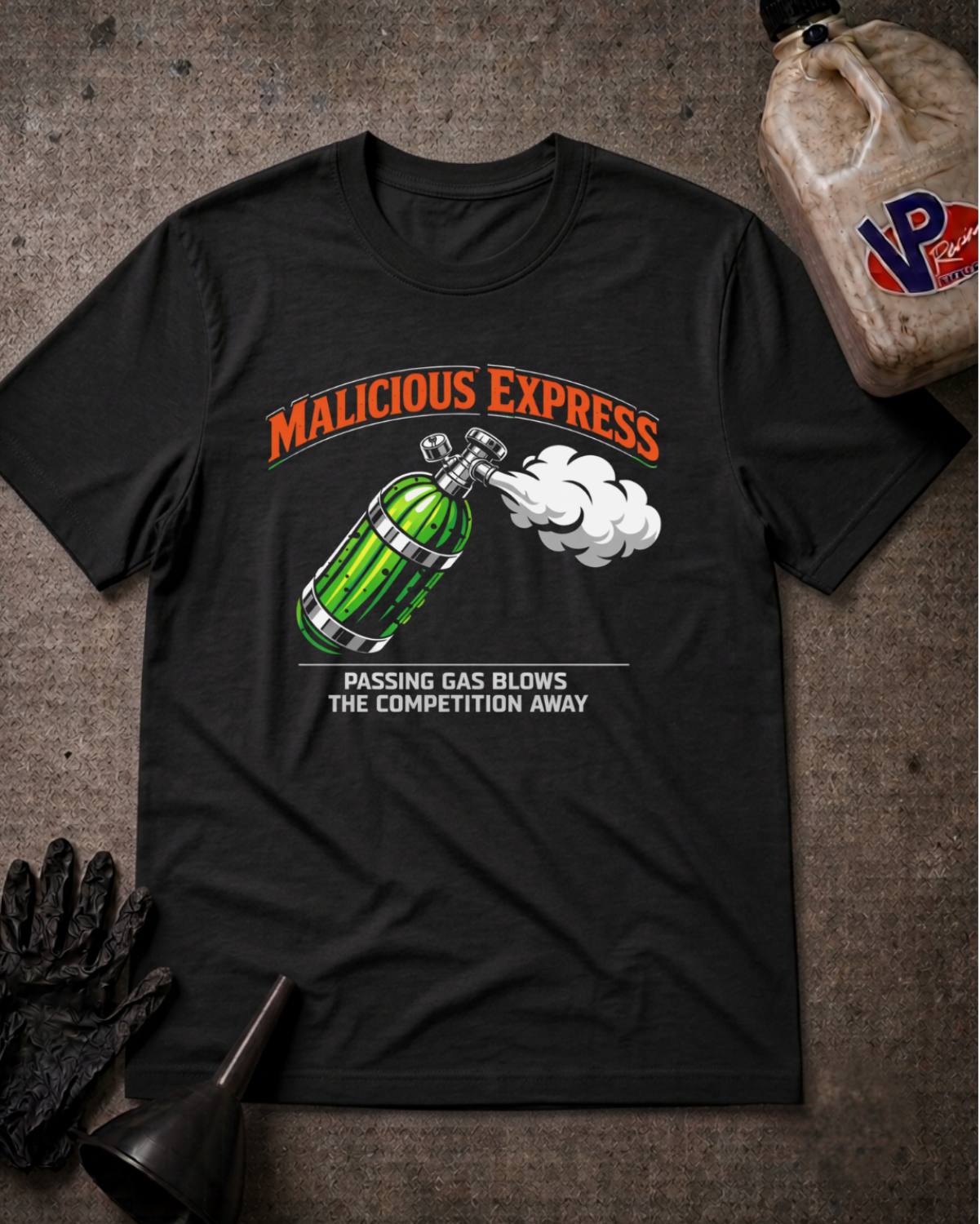 A black graphic t-shirt laid flat on a textured gray surface, surrounded by motorcycle-themed props including a black glove, a funnel, and a plastic jug. The shirt features a graphic of a pickle shaped nitrous bottle and 'MALICIOUS EXPRESS PASSING GAS BLOWS THE COMPETITION AWAY' text