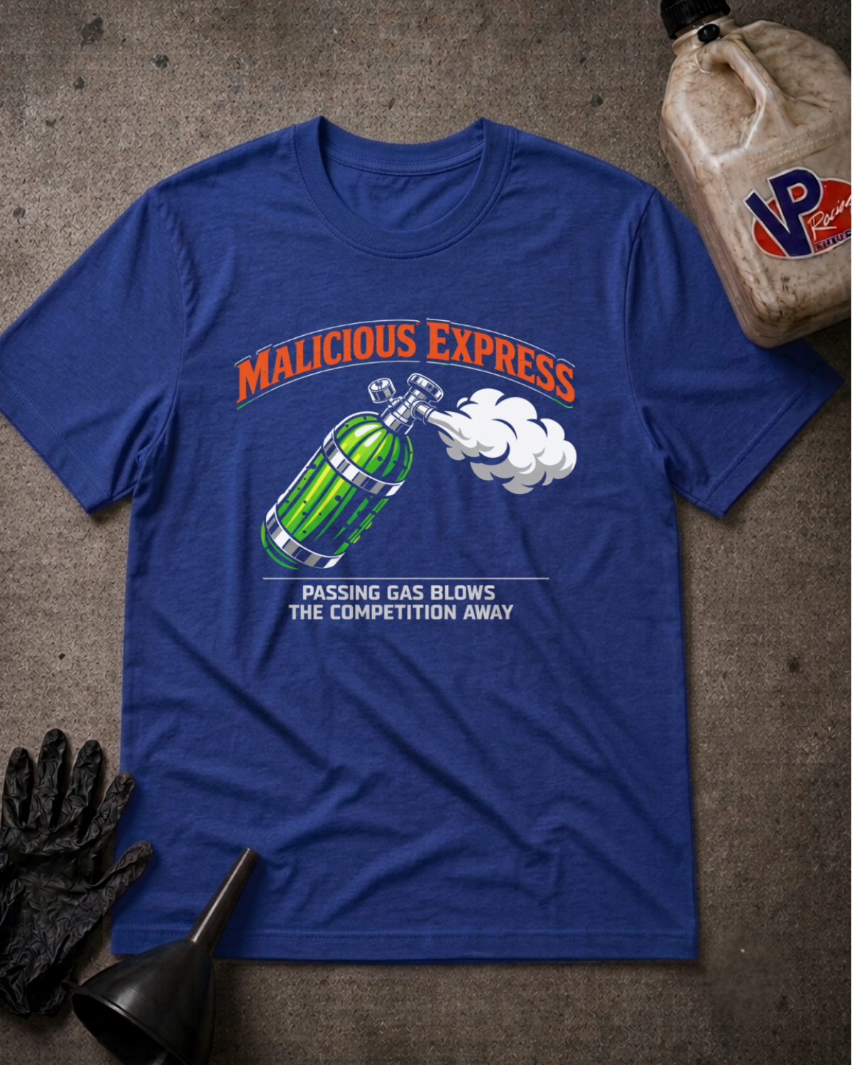 A royal blue graphic t-shirt laid flat on a textured gray concrete surface, surrounded by racing-themed props including an oil bottle, a black glove, a funnel. The t-shirt features a graphic of a pickle shaped nitrous bottle, with the text 'MALICIOUS EXPRESS' and 'PASSING GAS BLOWS THE COMPETITION AWAY'