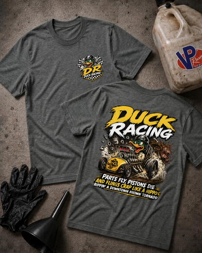 Two dark heather gray t-shirts displayed flat on a textured gray surface, showing the front and back of a 'Duck Racing' graphic design. The front features a small yellow and green chest logo, while the back displays a large graphic with a duck, race car, and humorous text.