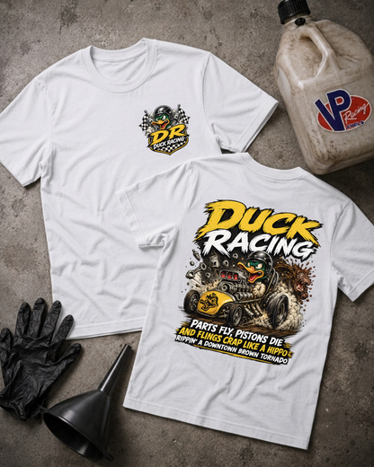 Two white graphic t-shirts displayed flat on a gray concrete surface, with a black funnel, black gloves, and a VP Racing fuel jug as props. The front shirt shows a small yellow and black chest logo, and the back shirt features a large 'Duck Racing' graphic with a race car and humorous text.